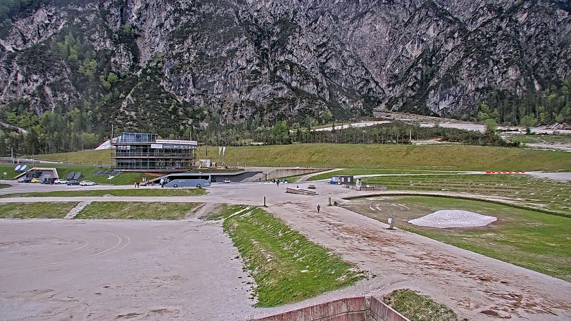 A large, gray building sits in a parking lot near a grassy area and a dirt track against a backdrop of a steep, gray mountain under an overcast sky.