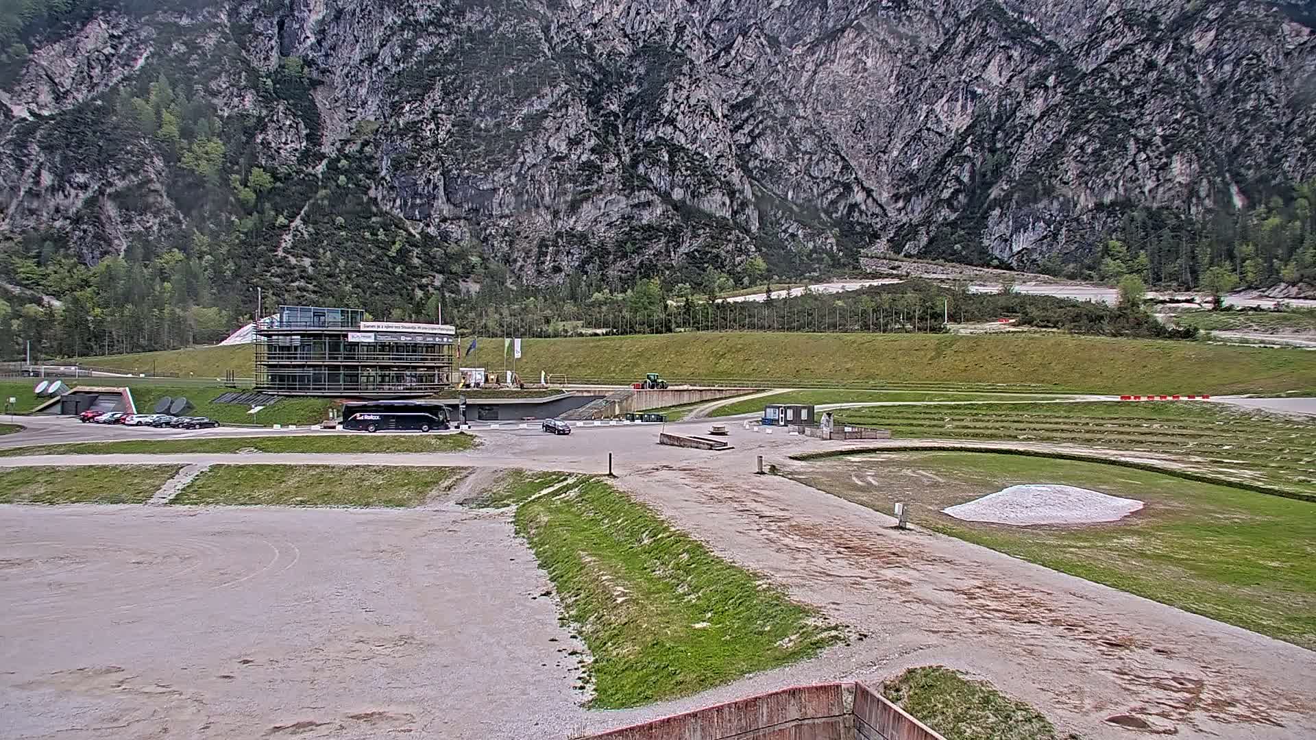A large, gray mountain overlooks a parking lot with several vehicles, a multi-story building, and a grassy area with what appears to be a small pile of light-colored material.
