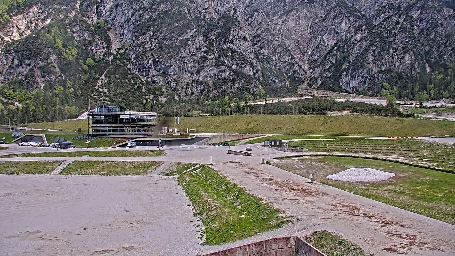 A modern building sits in a large, mostly empty, gravel parking lot at the base of a large, gray mountain on an overcast day.