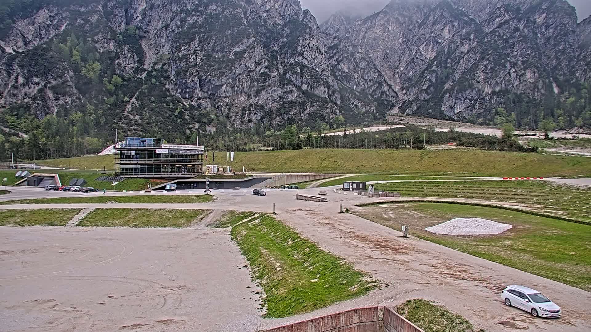 A modern building sits in a valley between large, grey mountains under an overcast sky, with a gravel area and several cars visible.