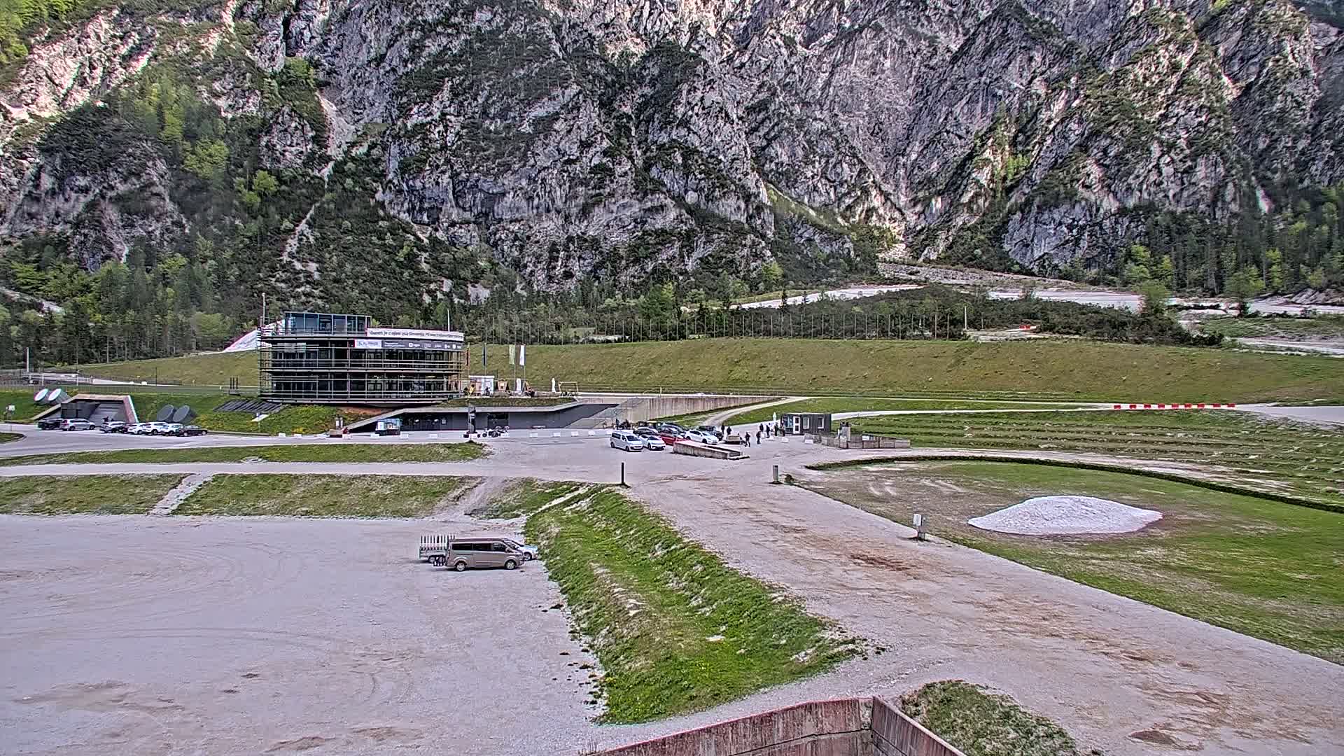 A large, modern building sits in a parking lot near a grassy area and track, all situated at the base of a large, rocky mountain under a cloudy sky.