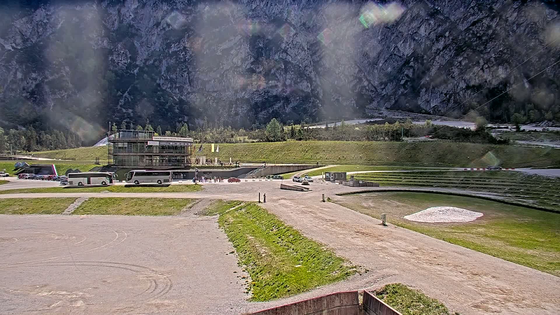A sunny day at a mountainside outdoor venue with several vehicles and buses parked near a building, overlooking a grassy area with tiered seating and a gravel track.