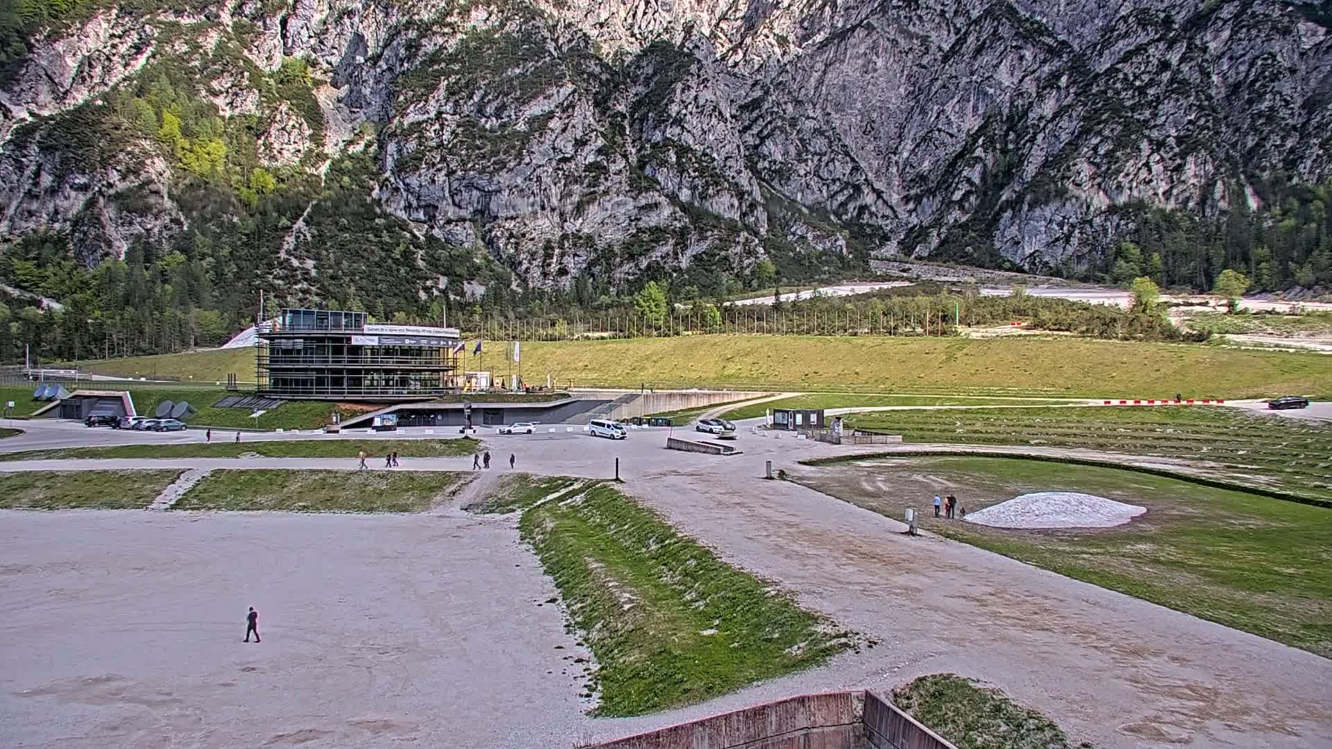 A modern building sits in a parking lot overlooking a grassy field and a large, rocky mountain under a partly cloudy sky.