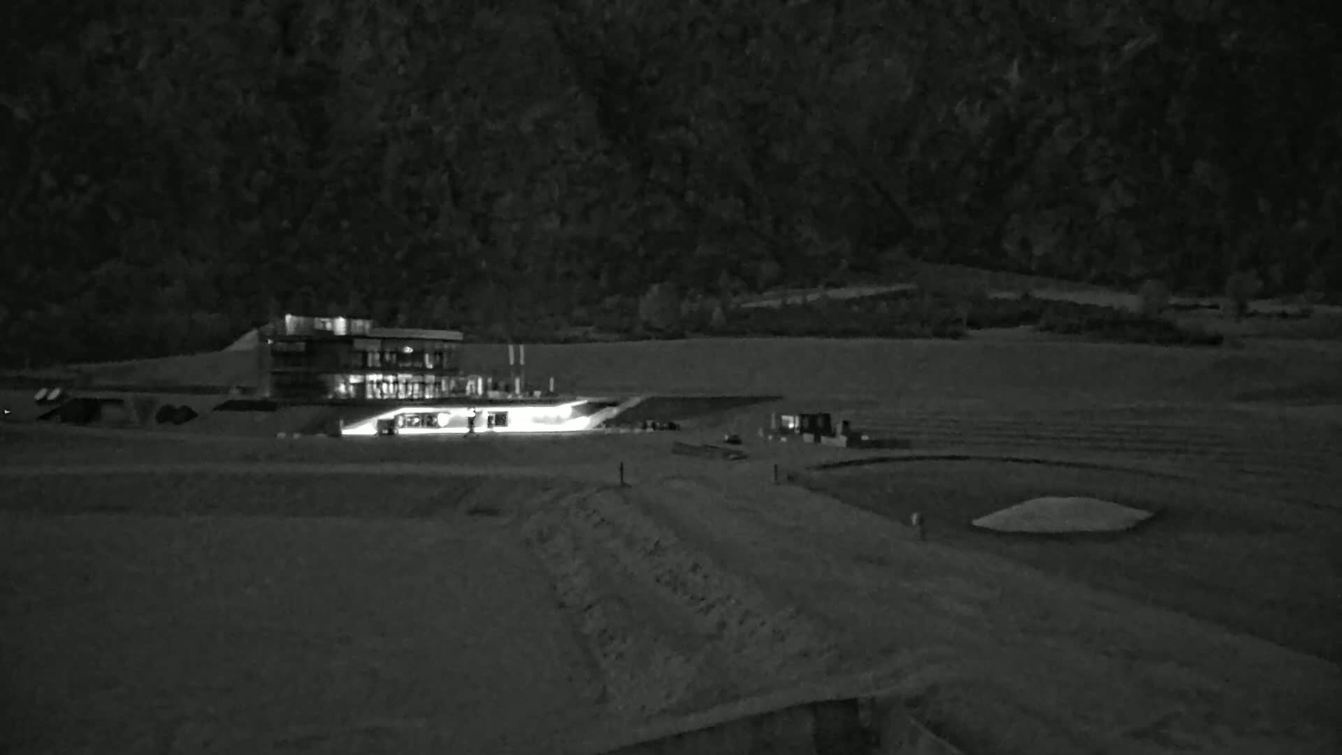 A lighted building sits on a mostly flat, grassy area at night, with a hilly, dark landscape in the background.