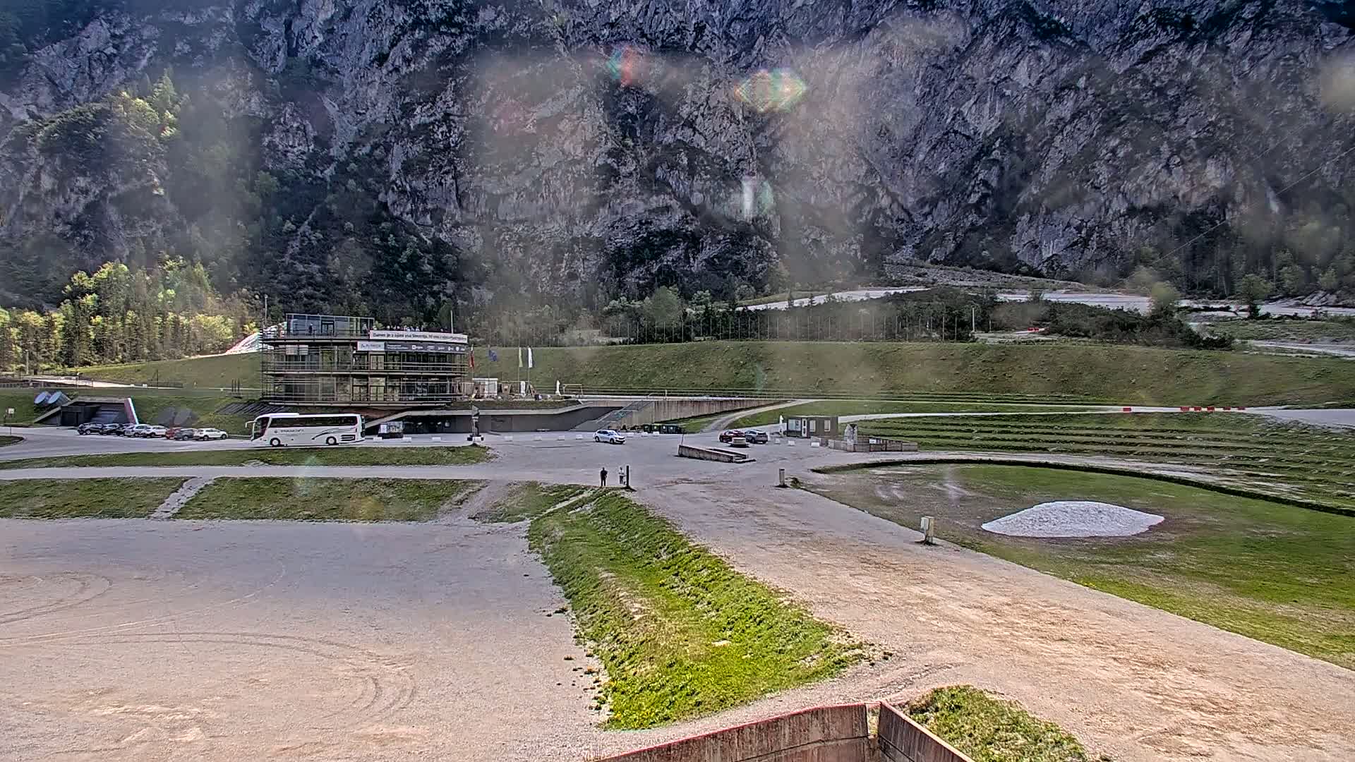 A large, multi-story building sits in a parking lot next to a grassy track at the base of a mountain range under an overcast sky.