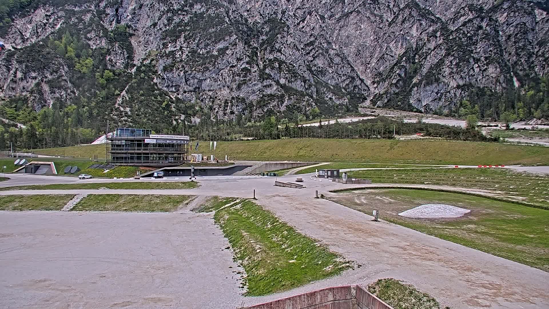 A mostly empty paved area with a multi-story building under construction sits in front of a large, rocky mountain under an overcast sky.