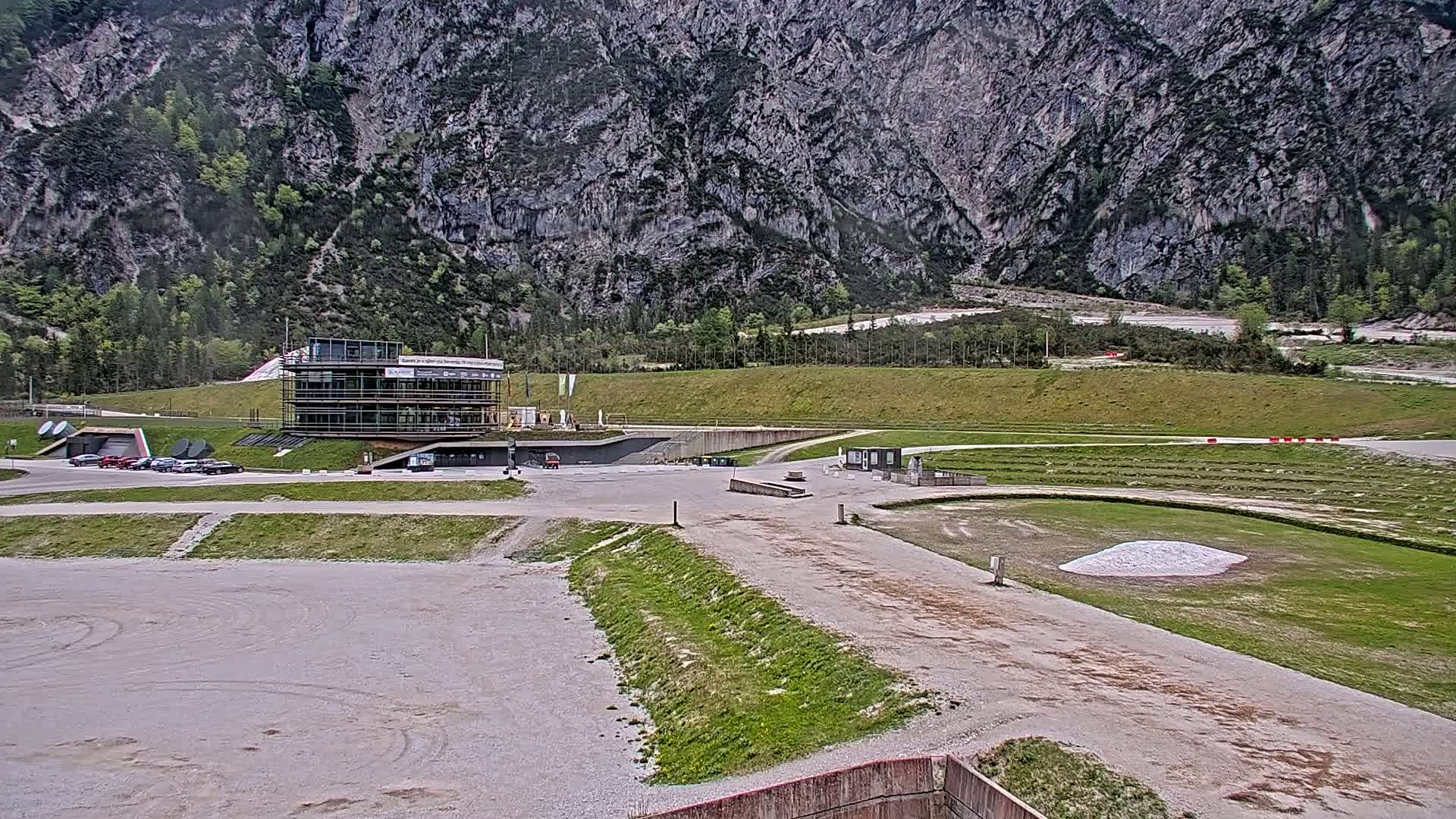 A modern building sits in a large, mostly empty parking lot overlooking a grassy field and a rocky mountainside under an overcast sky.
