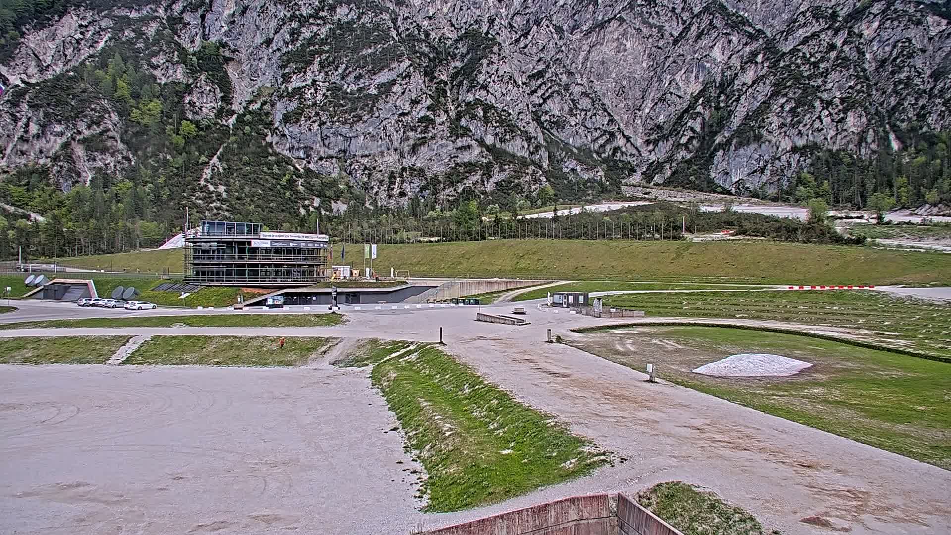 A modern building sits in a large, mostly empty paved area before a grassy field and a steep, gray mountain under an overcast sky.