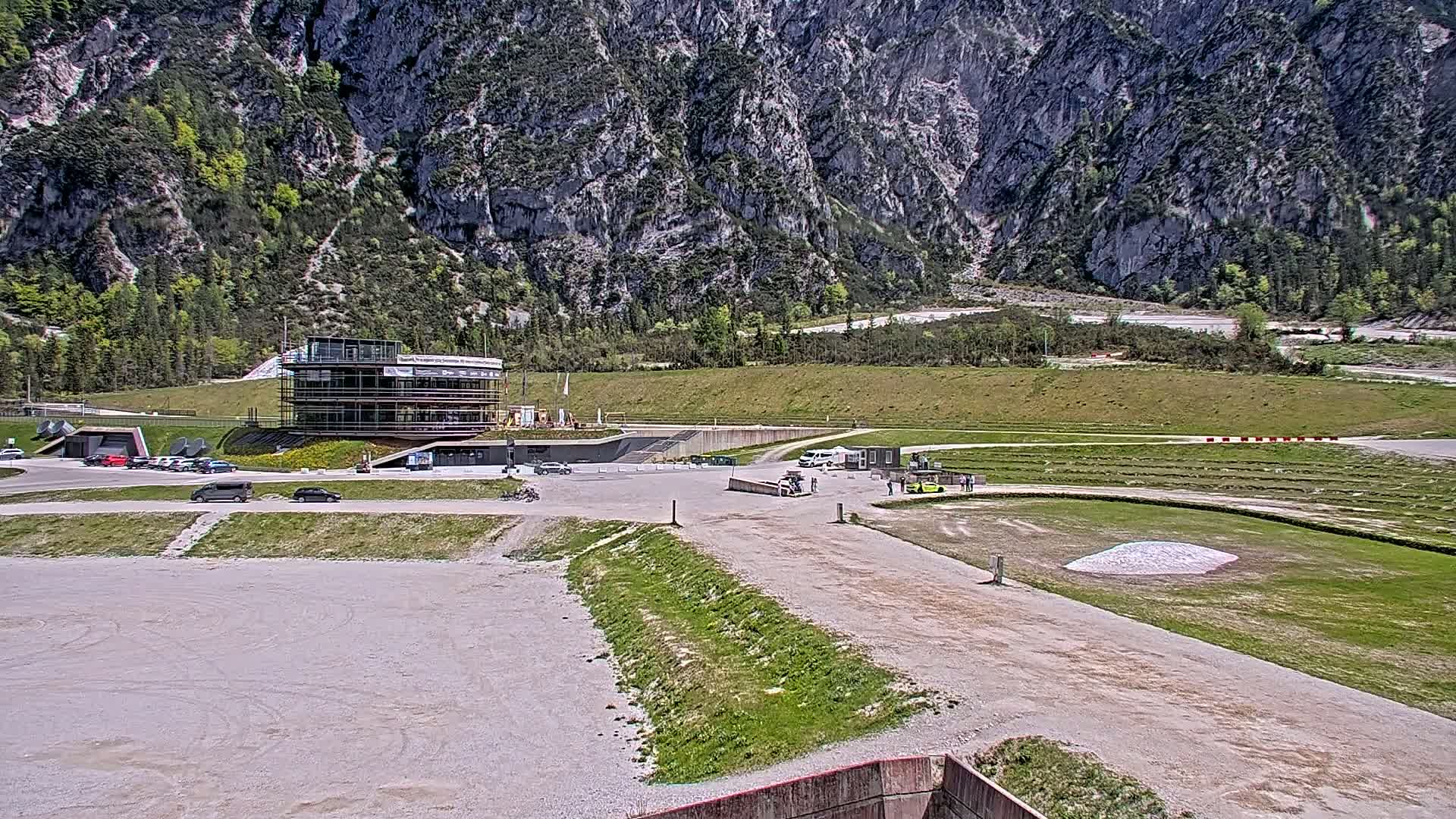 A multi-story building sits in a parking lot next to a grassy area and track, all nestled in a valley with a large mountain in the background on a sunny day.