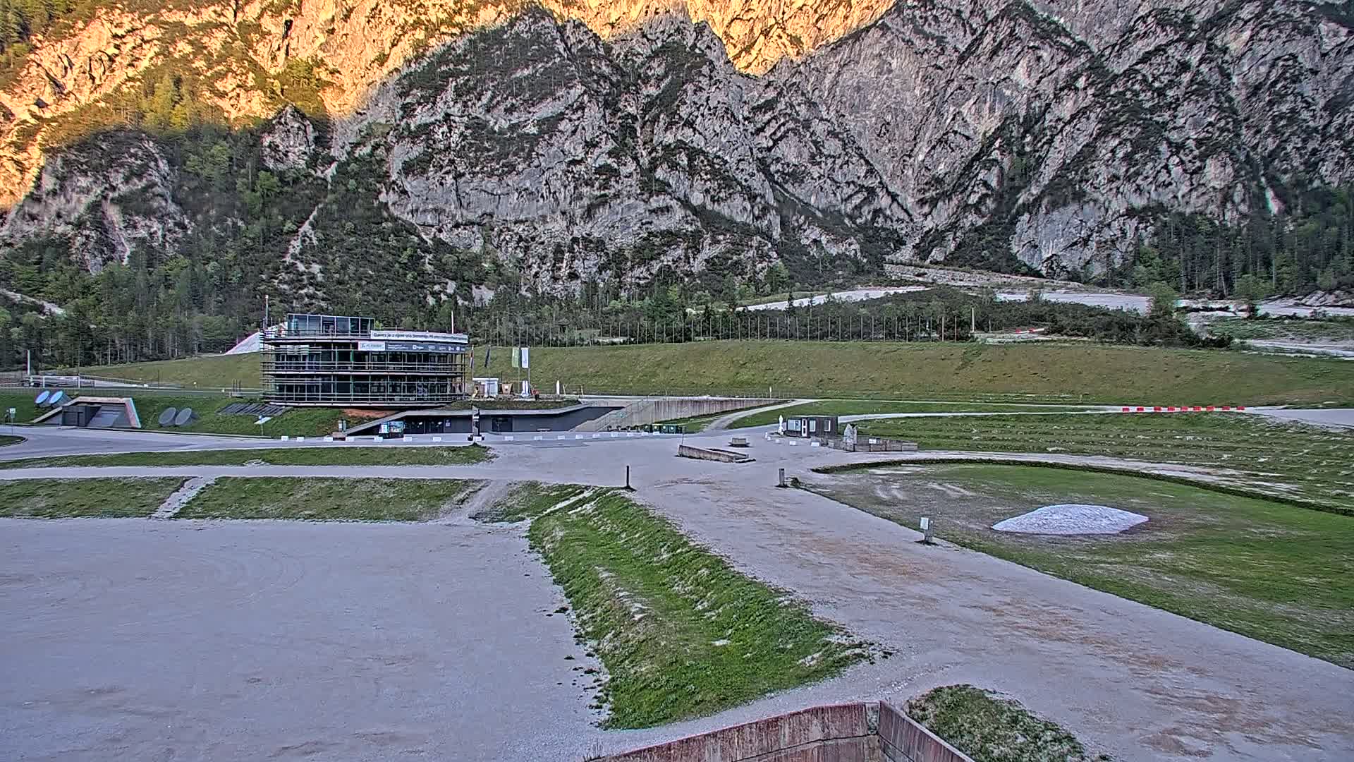 A multi-story building sits in a large, mostly empty, outdoor area with patches of grass, against a backdrop of towering, light-brown, rocky mountains under a clear sky.