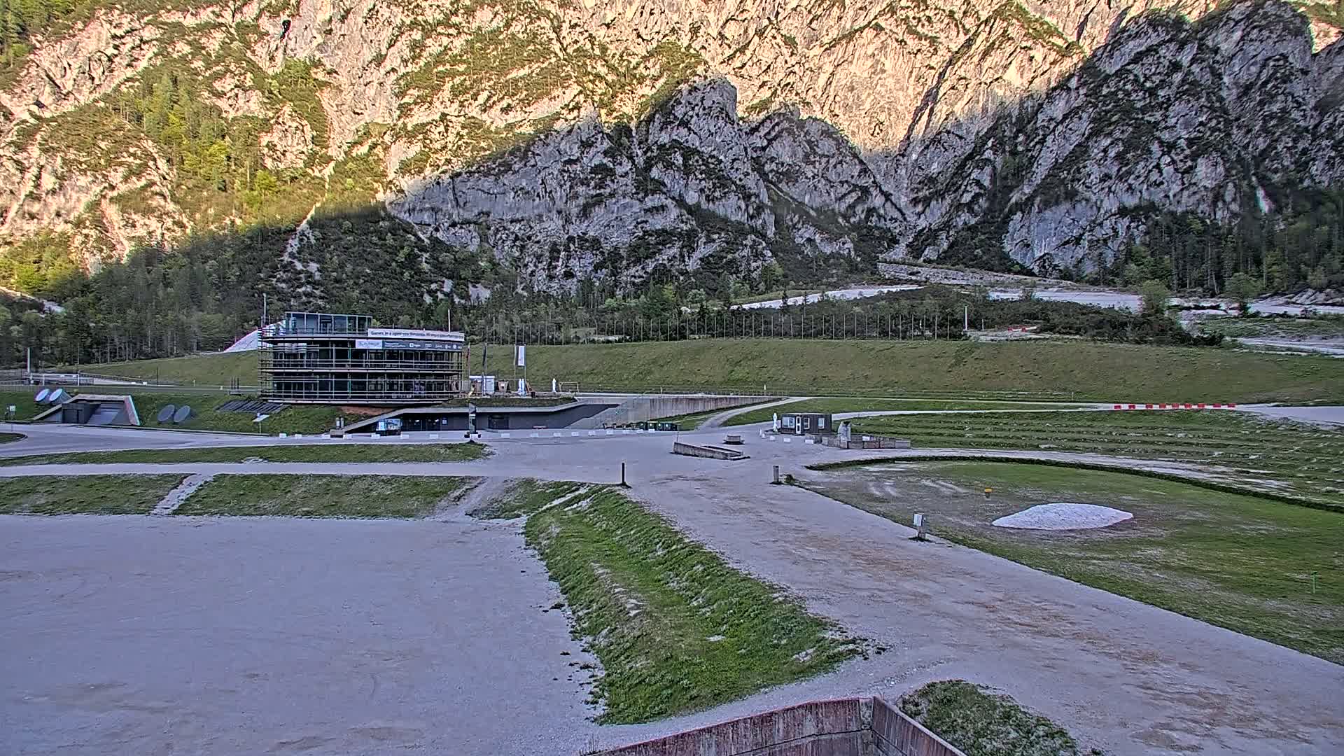 A two-story building sits in a large, mostly empty outdoor area near a tall, rocky mountain under a partly sunny sky.