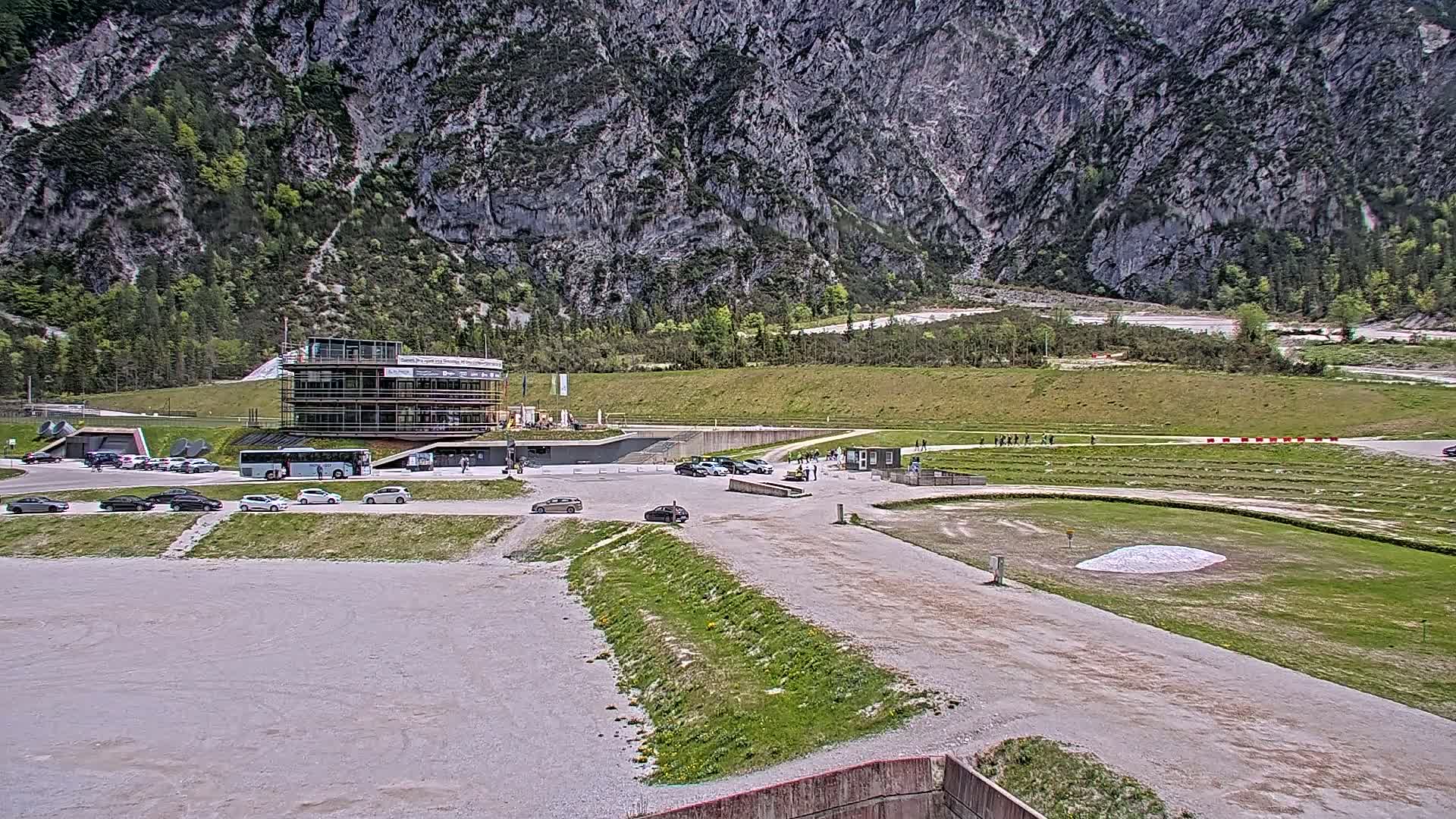 A modern building and parking lot are situated in a valley, with a large, rocky mountain in the background under a partly cloudy sky.