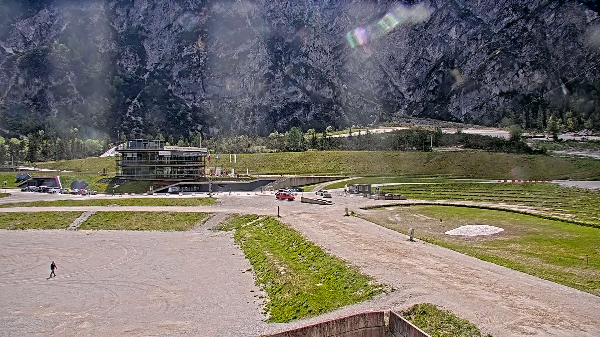 A modern building sits at the base of a large, rocky mountain on a sunny day, with a gravel area and a grassy field in the foreground.