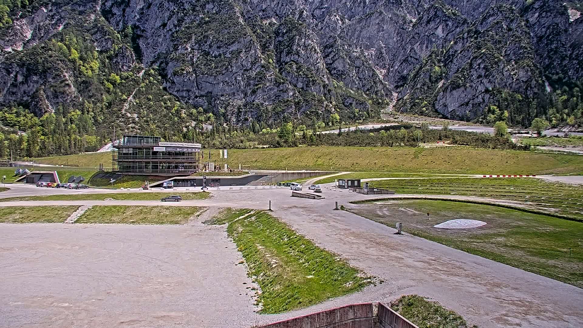 A parking lot and modern building are situated in a valley surrounded by rocky mountains under a bright, sunny sky.