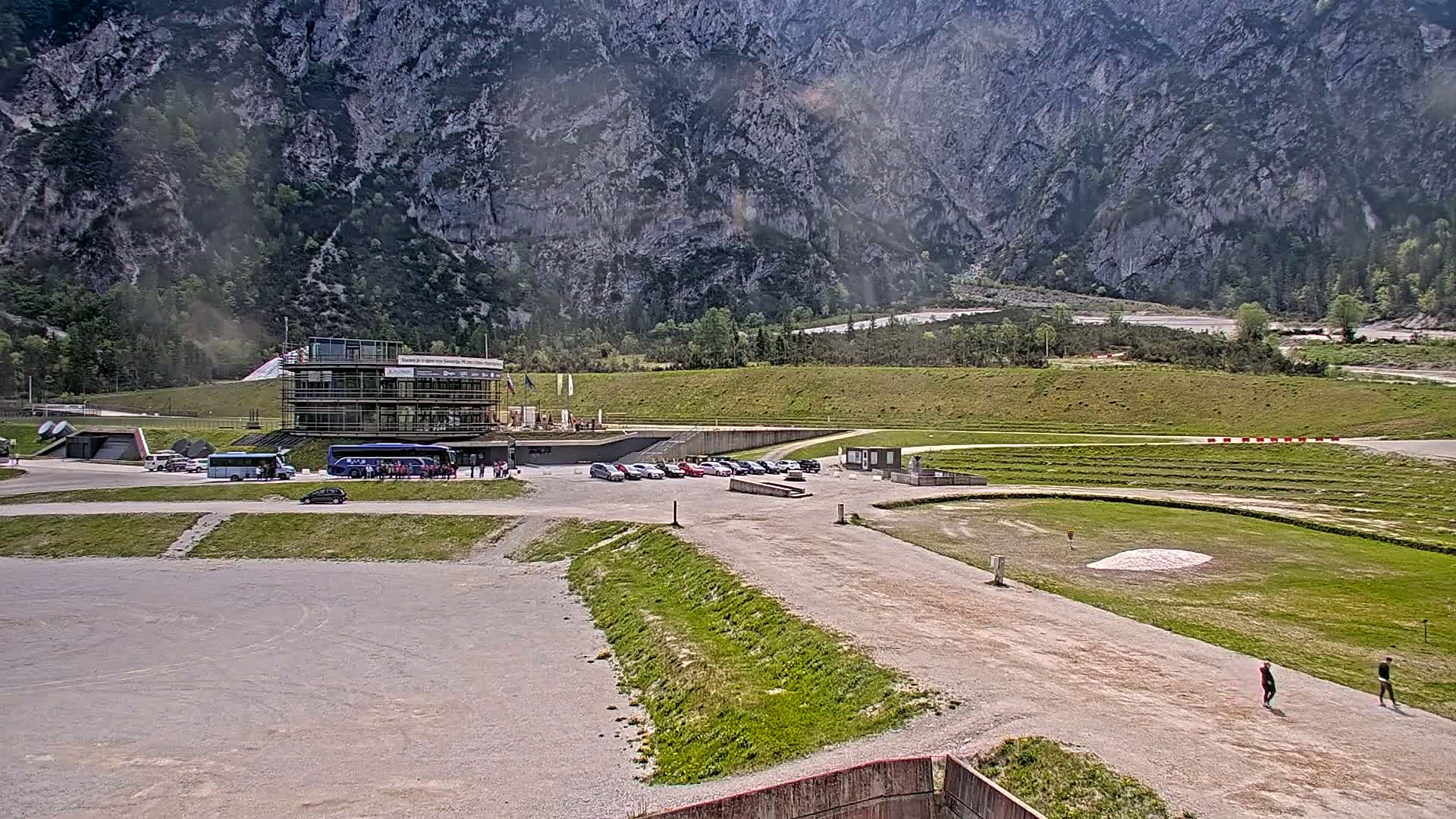 A paved area with a building, parked cars, and a few people, situated in front of a large, rocky mountain under a sunny sky.