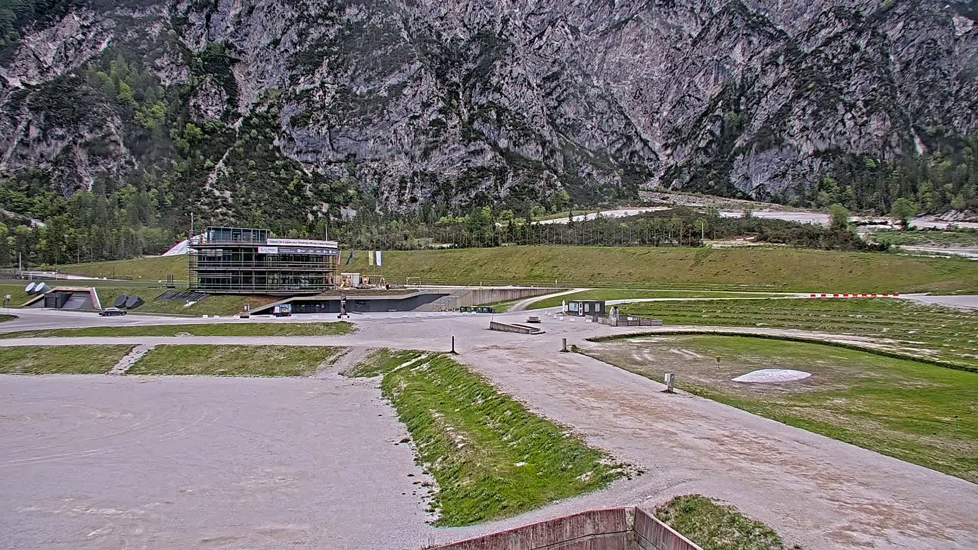 A modern building sits in a large, mostly empty paved area at the base of a large, rocky mountain under an overcast sky.