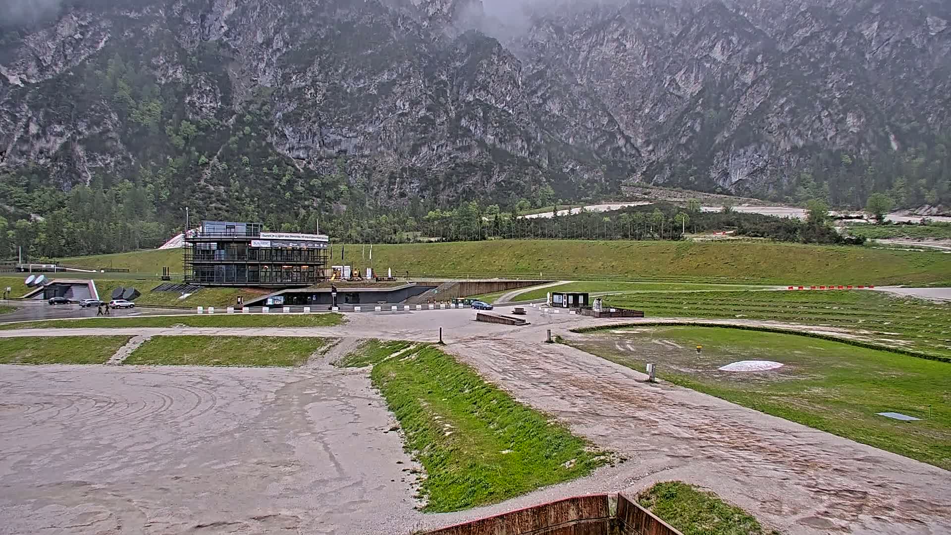 A modern building sits in a gravel parking lot overlooking a grassy field and a mountain range under an overcast sky.