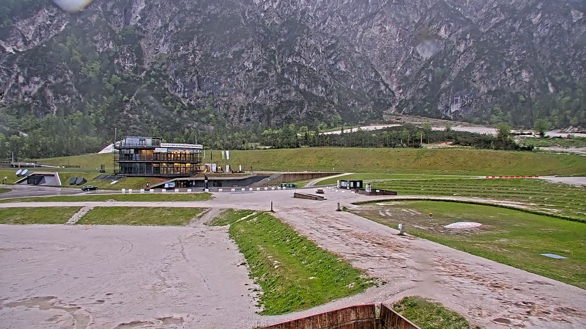 A modern building sits at the base of a large, gray mountain range on an overcast day, overlooking a flat, grassy area with a dirt track.