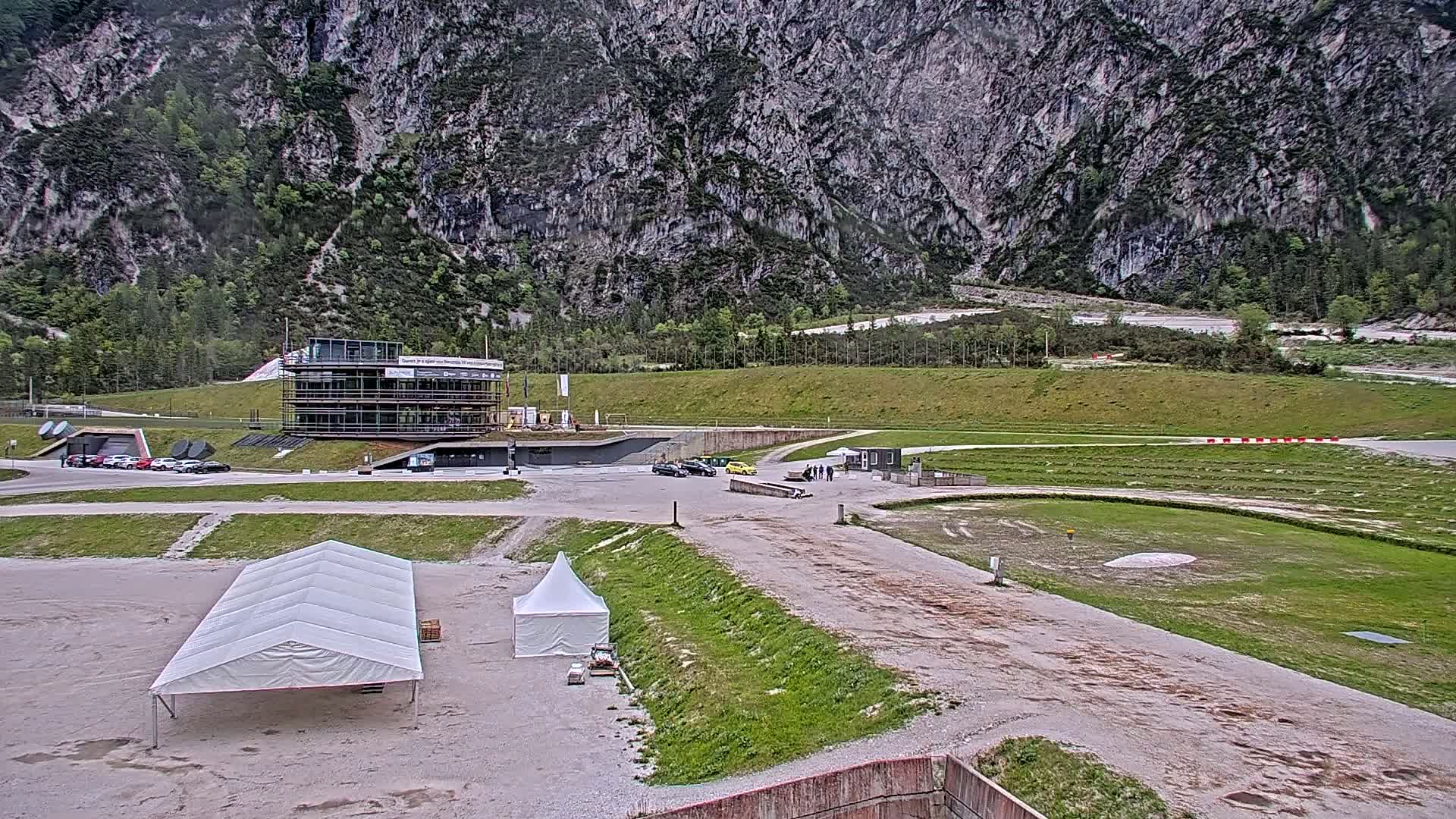 A mostly overcast day reveals a mountainside view of a sports facility with several cars parked near a building, several white tents, and a grassy area in the foreground.