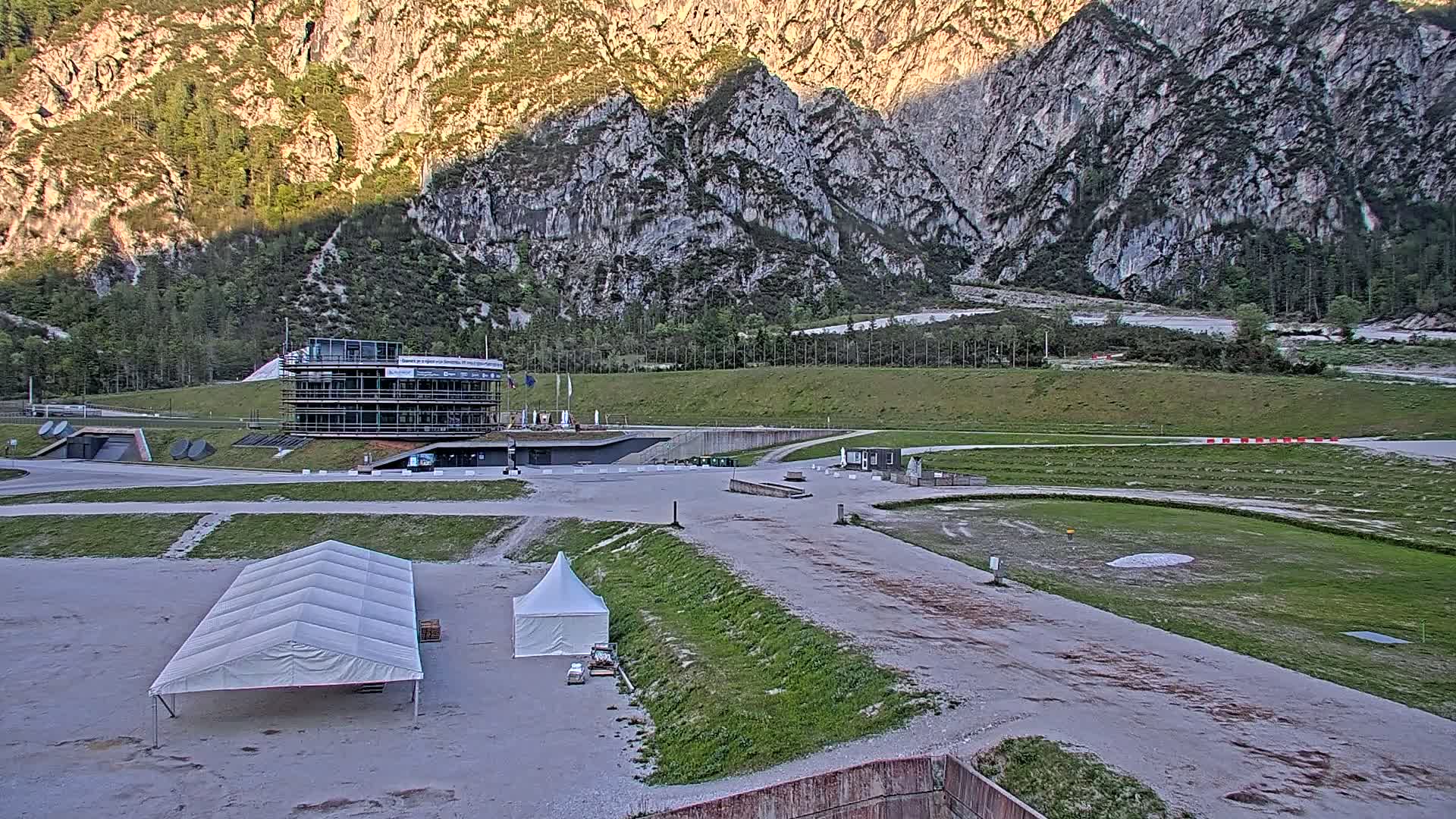 A large building sits in a wide, open area next to several white tents, with a grassy field and rocky mountains in the background under a clear sky.