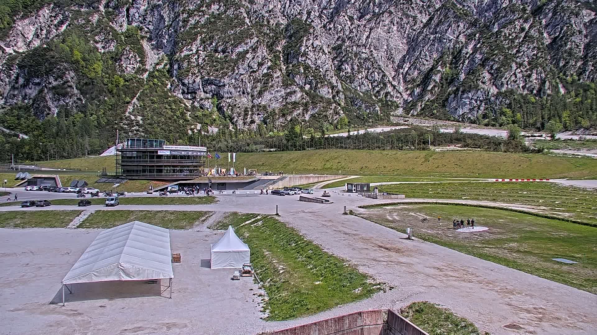 A large building and several tents sit in a parking lot at the base of a rocky mountain on a sunny day.