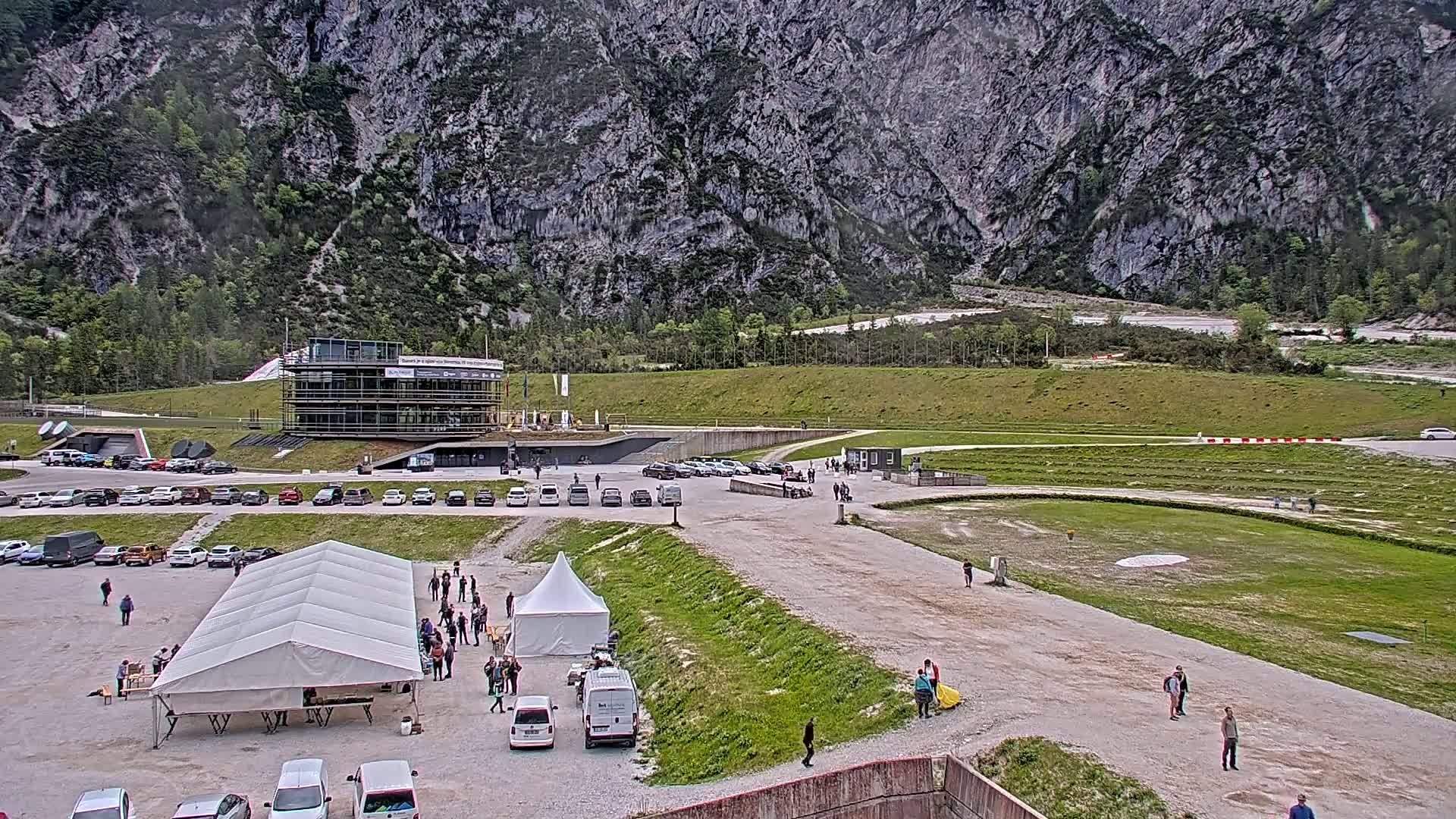 A large, mostly empty parking lot with several tents and people, situated in front of a modern building and a grassy area against a backdrop of a large, gray mountain under an overcast sky.
