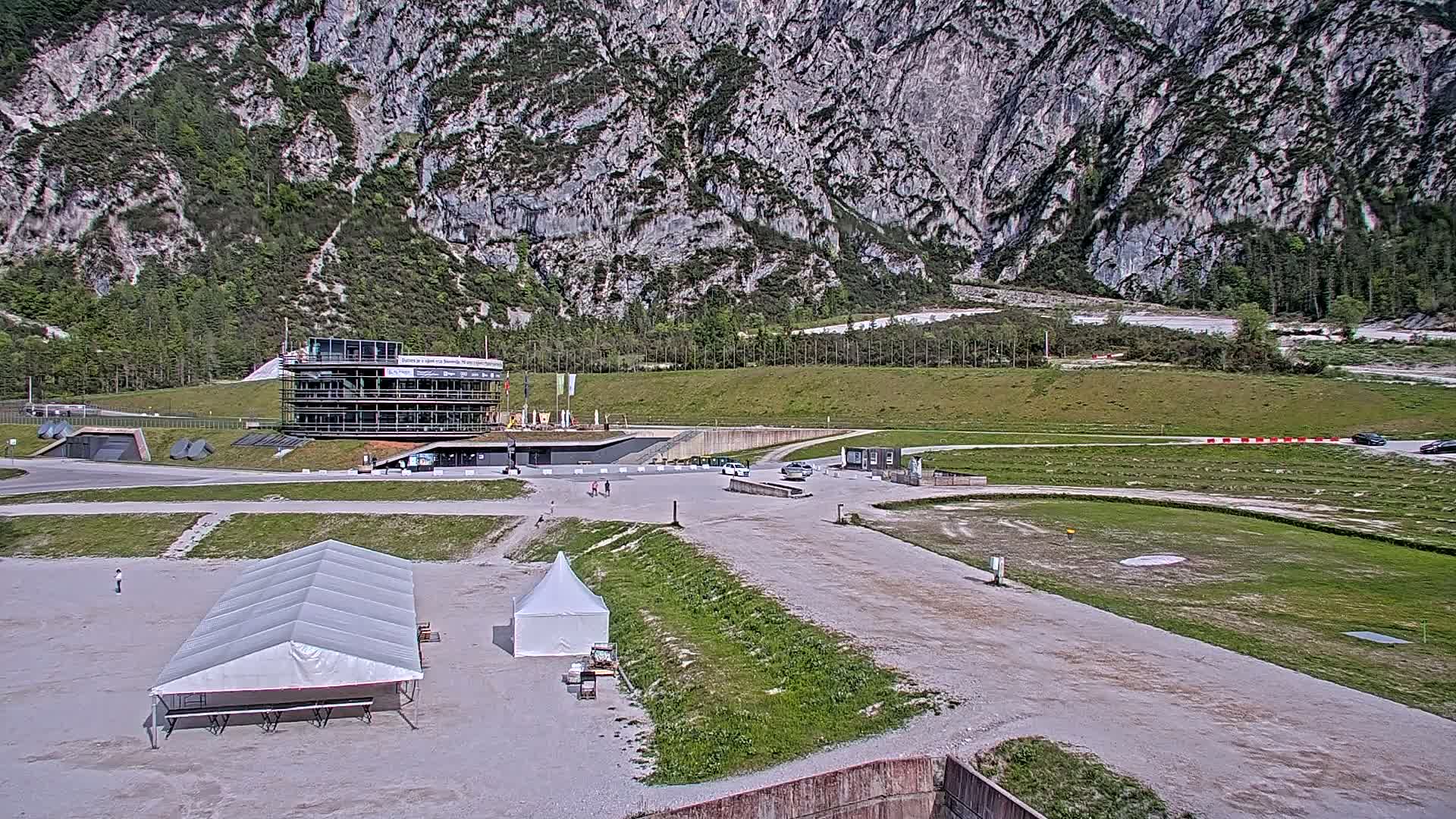 A large building complex sits in a flat area at the base of a large, rocky mountain on a sunny day, with several small tents and a few cars visible in a parking lot.