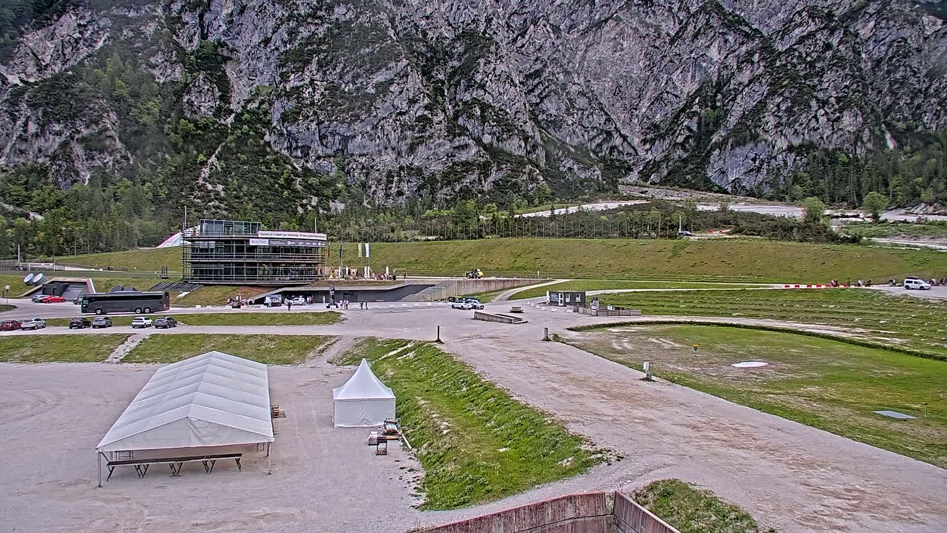 A large, multi-story building sits near a grassy field and parking lot in front of a rocky mountain under an overcast sky.