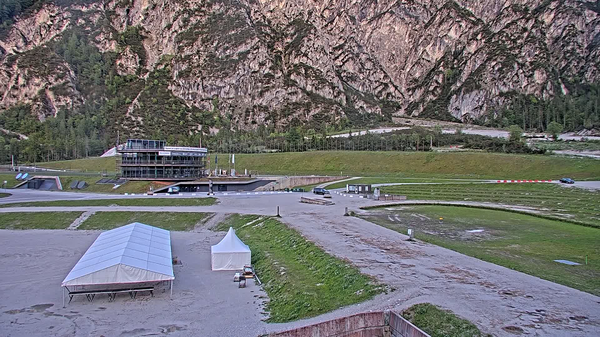 A large building sits in a flat area near a mountain, with two white tents and a vehicle in the parking lot, under overcast skies.