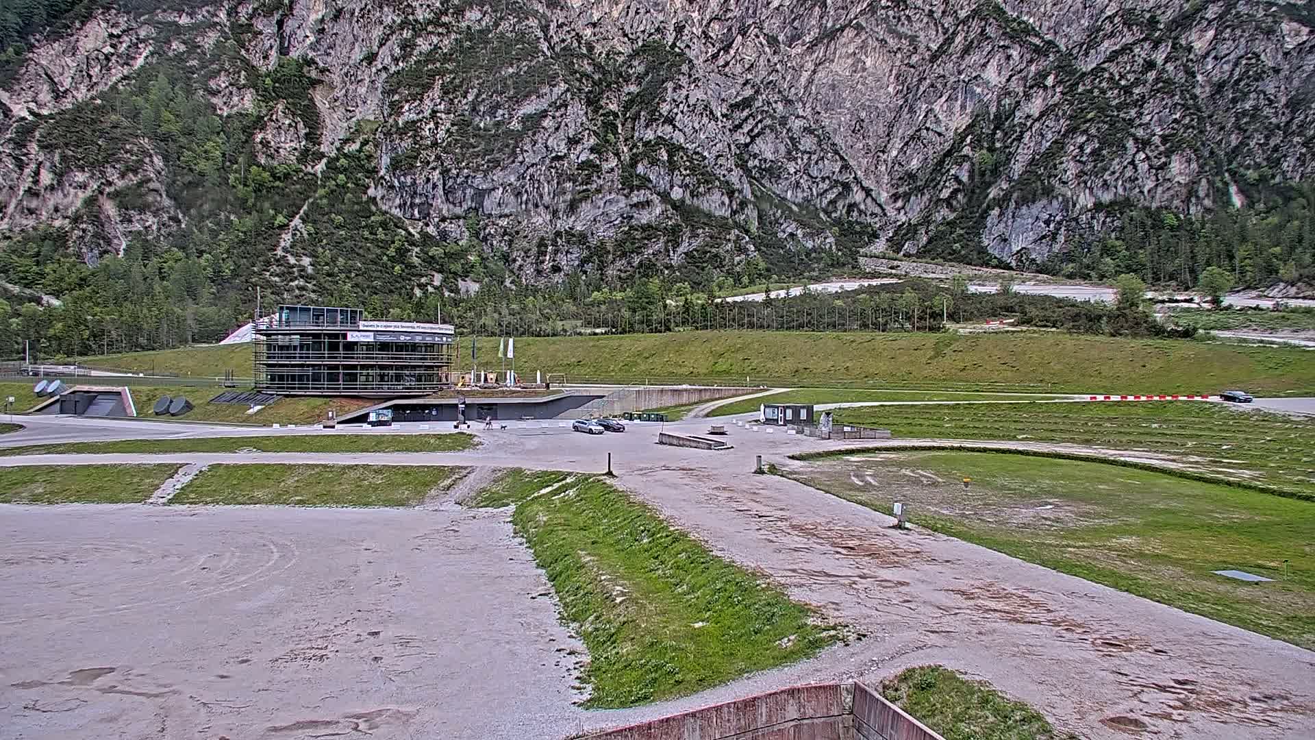 A large, two-story building sits in a parking lot at the base of a rocky mountain on a partly cloudy day.