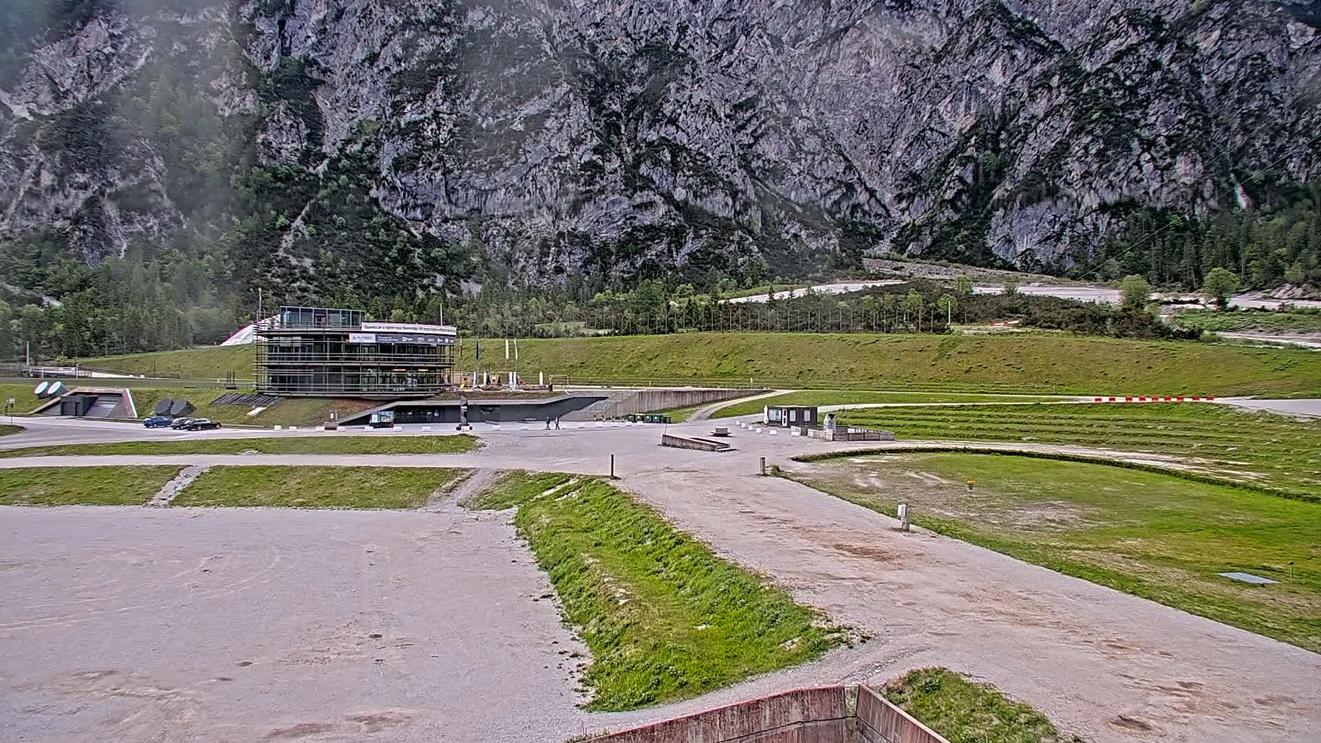 A modern building sits in a large, mostly empty paved area next to a grassy field and track against a backdrop of a large, gray, rocky mountain under an overcast sky.