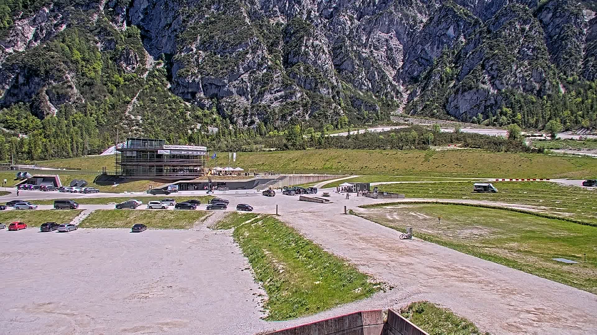 A parking lot with cars sits in front of a multi-story building under construction, near a grassy area and track against a backdrop of a large, rocky mountain on a sunny day.