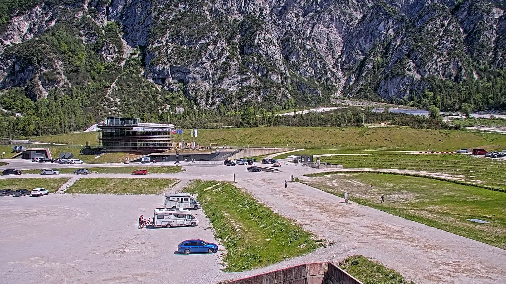 A parking lot with several cars and RVs is situated in front of a multi-story building near a grassy field and a large, rocky mountain under a sunny sky.
