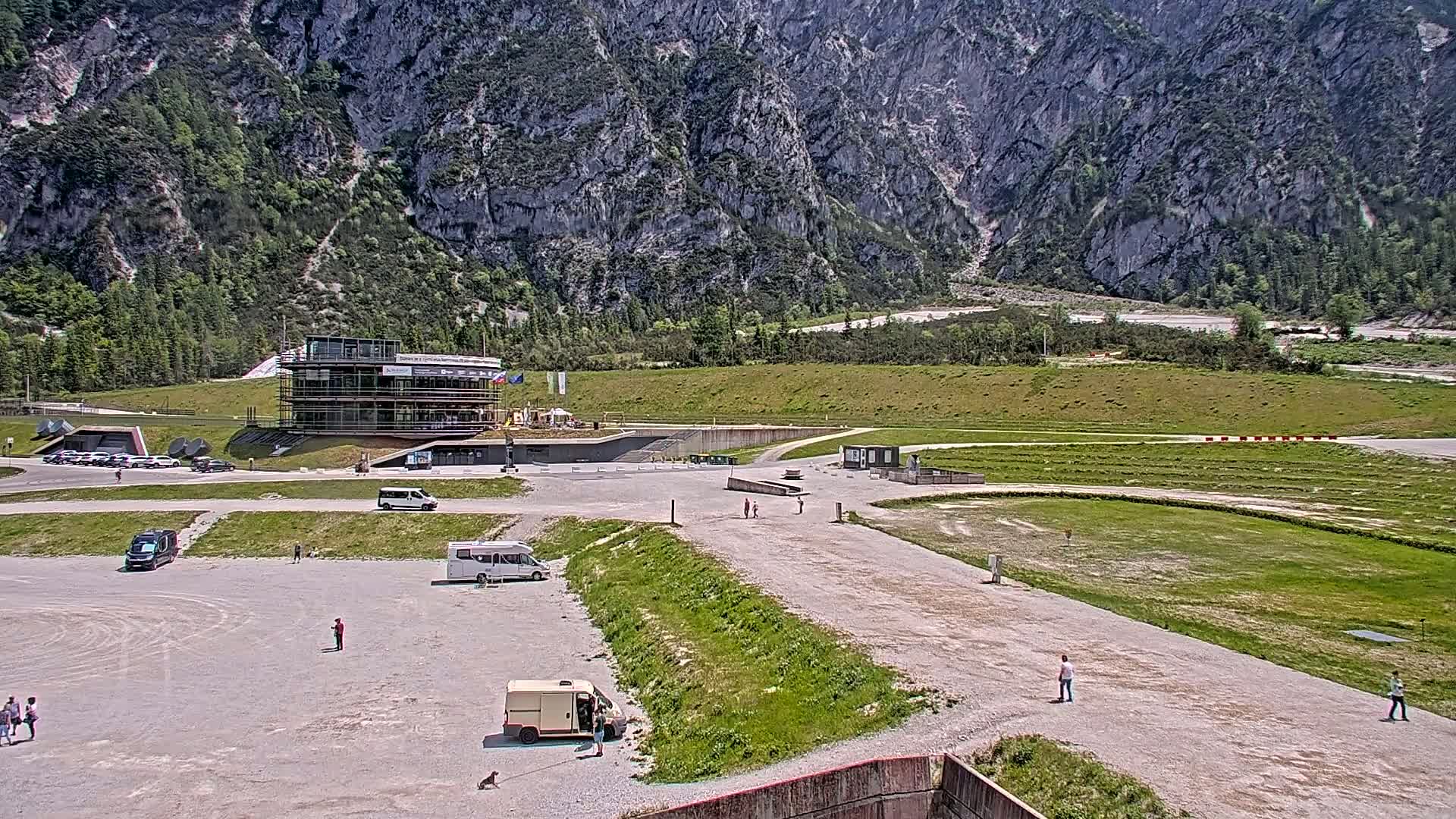 A large, mostly empty parking lot and grassy areas sit before a multi-story building under construction, all situated against a backdrop of a mountain range on a sunny day.