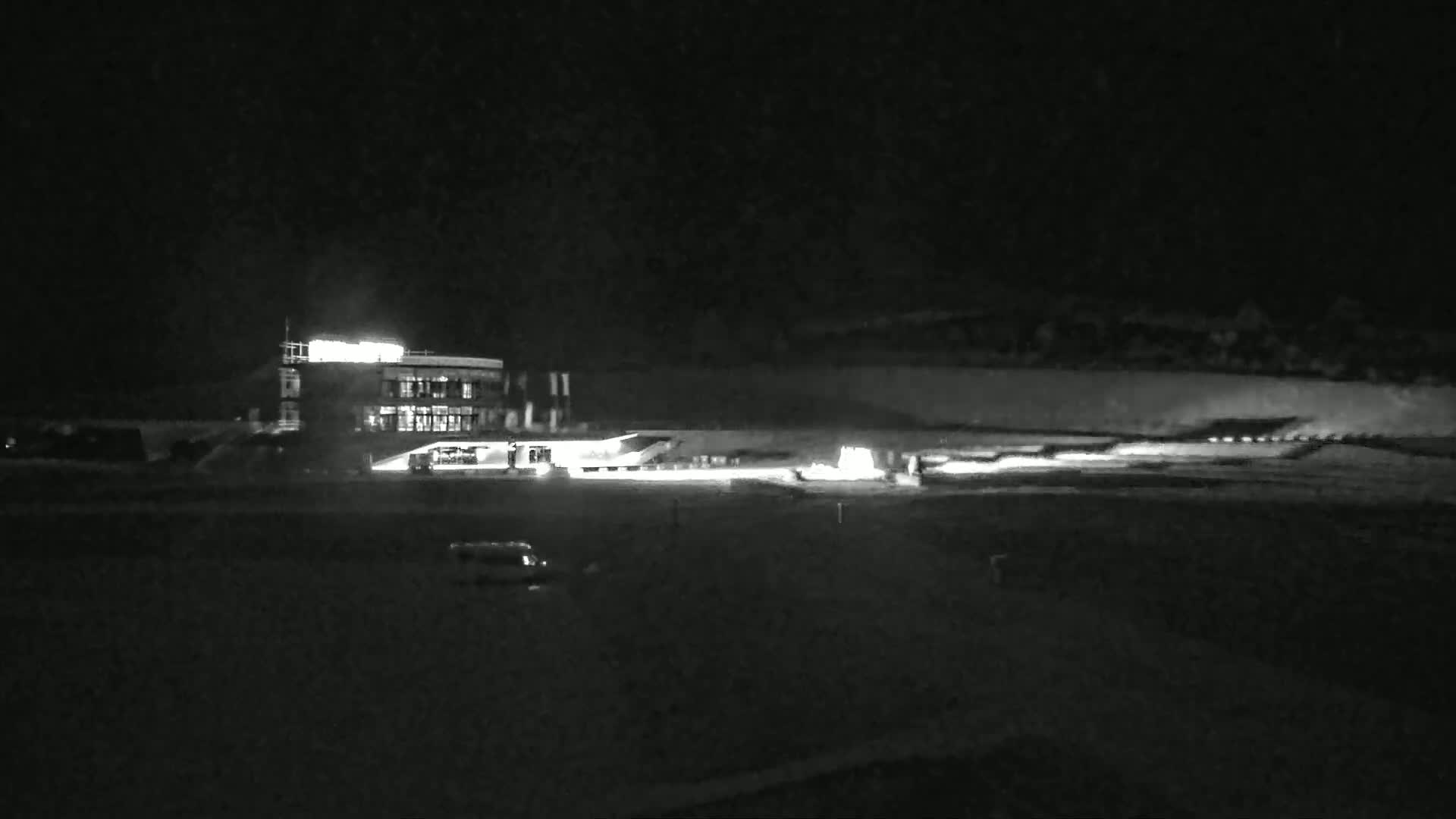 A brightly lit building is visible at night, with a roadway and some vehicles in front of it under dark, clear skies.