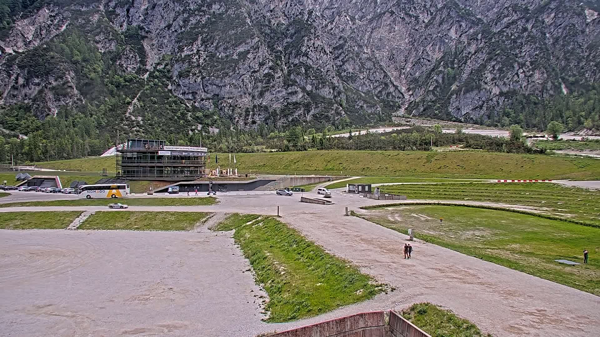 A modern building complex sits in a valley before a large, grey mountain under an overcast sky.
