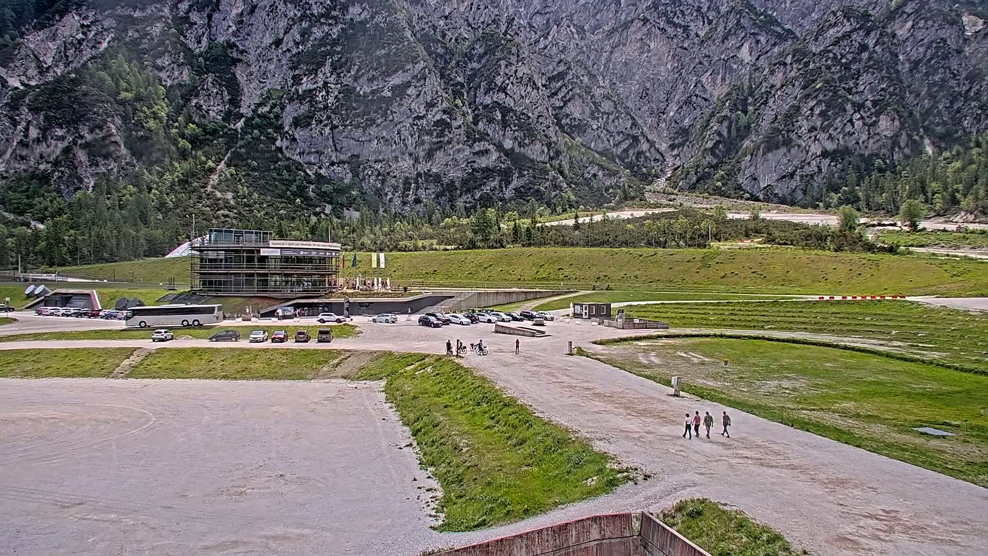 A multi-story building with a parking lot and several people walking sits in front of a large, grey mountain under a partly cloudy sky.