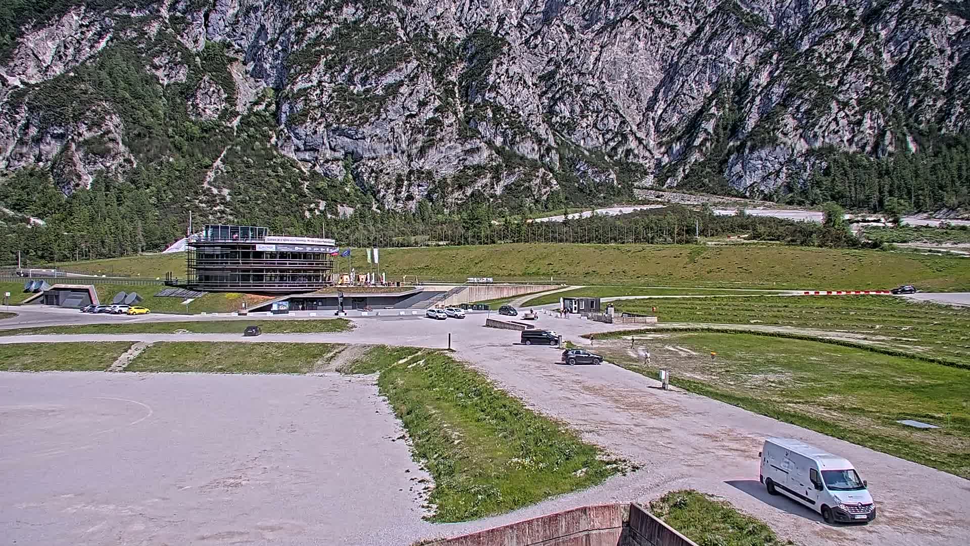 A multi-story building sits in a parking lot next to a grassy area and racetrack, all set against a backdrop of a large, rocky mountain on a sunny day.