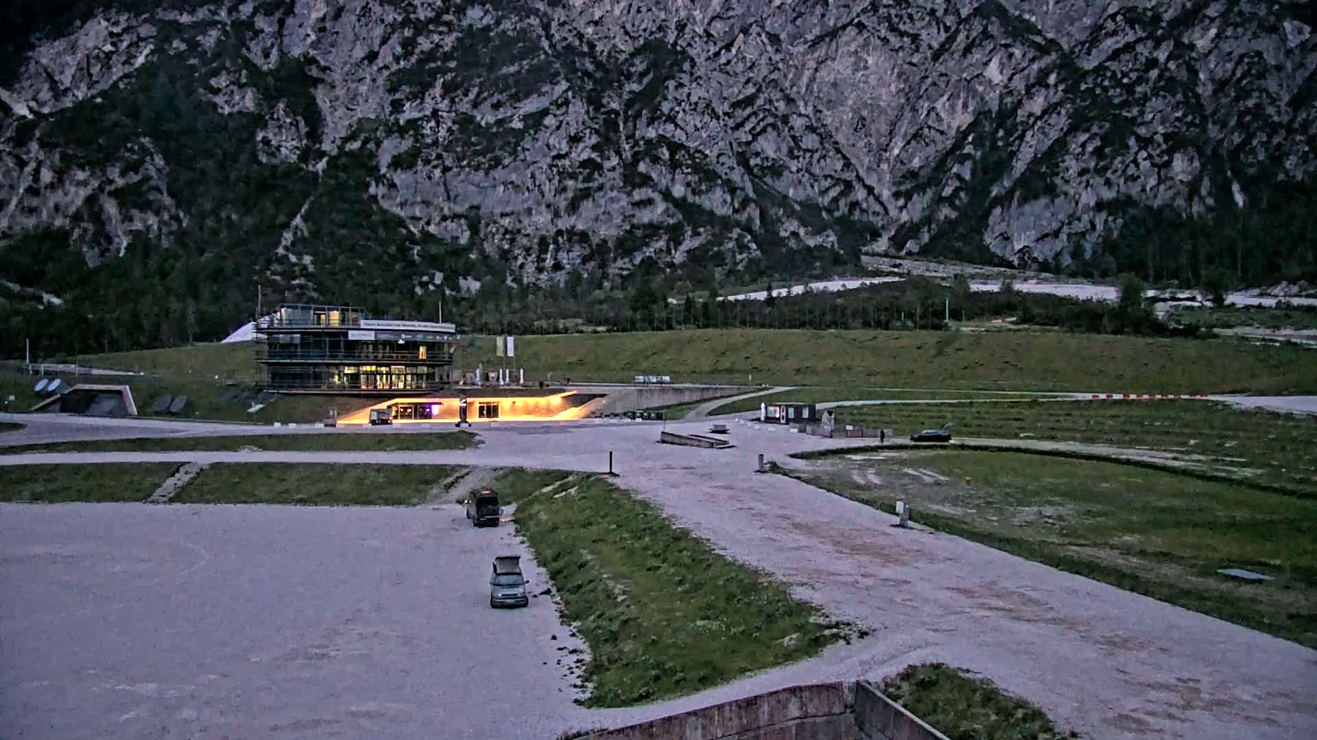 A building complex sits in a large, flat, light-grey area next to a grassy field, all set against a backdrop of a large, grey, mountainous landscape under a twilight sky.