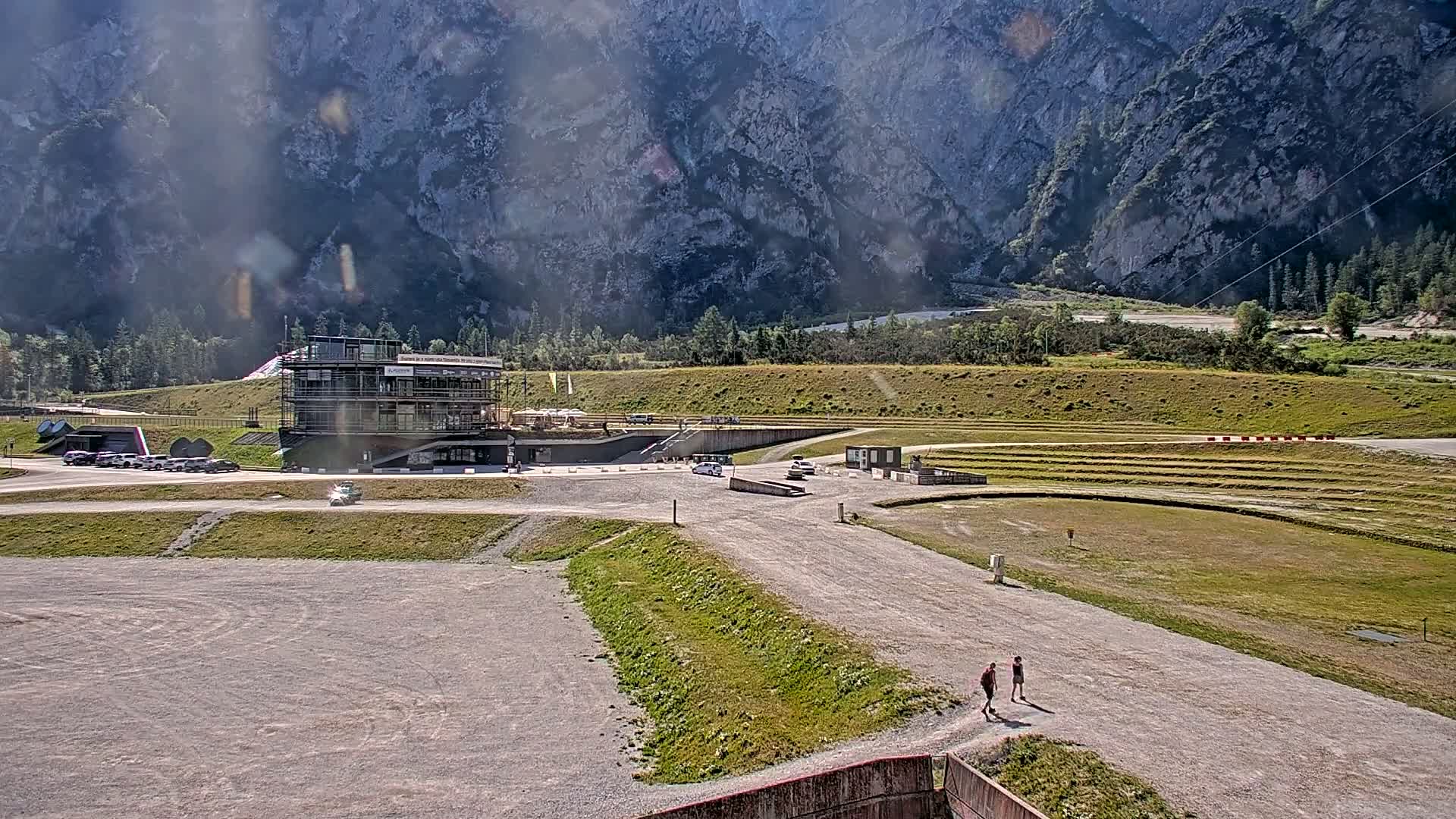 A gravelly area with a multi-story building and a few cars, two people walking, and a large grassy area sits before a mountainous backdrop on a sunny day.
