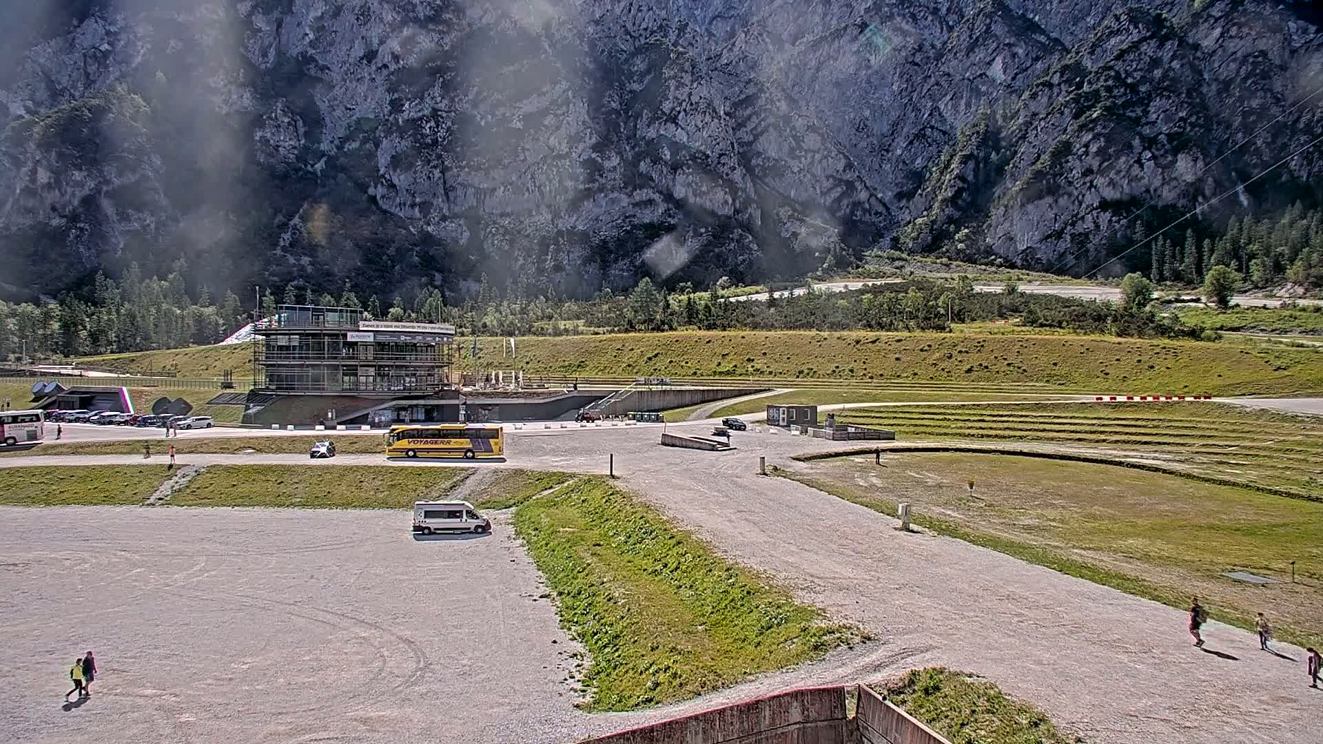 A large, open gravel area with a few vehicles and pedestrians, a multi-story building, and grassy areas sits in front of a large, rocky mountain under a sunny sky.