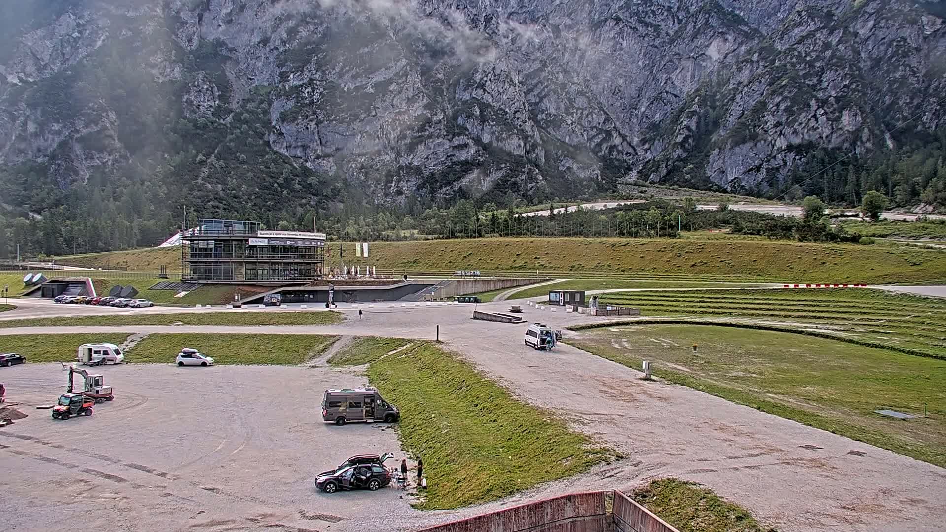 A parking lot with several vehicles and a modern building is situated in front of a large, rocky mountain under a cloudy sky.