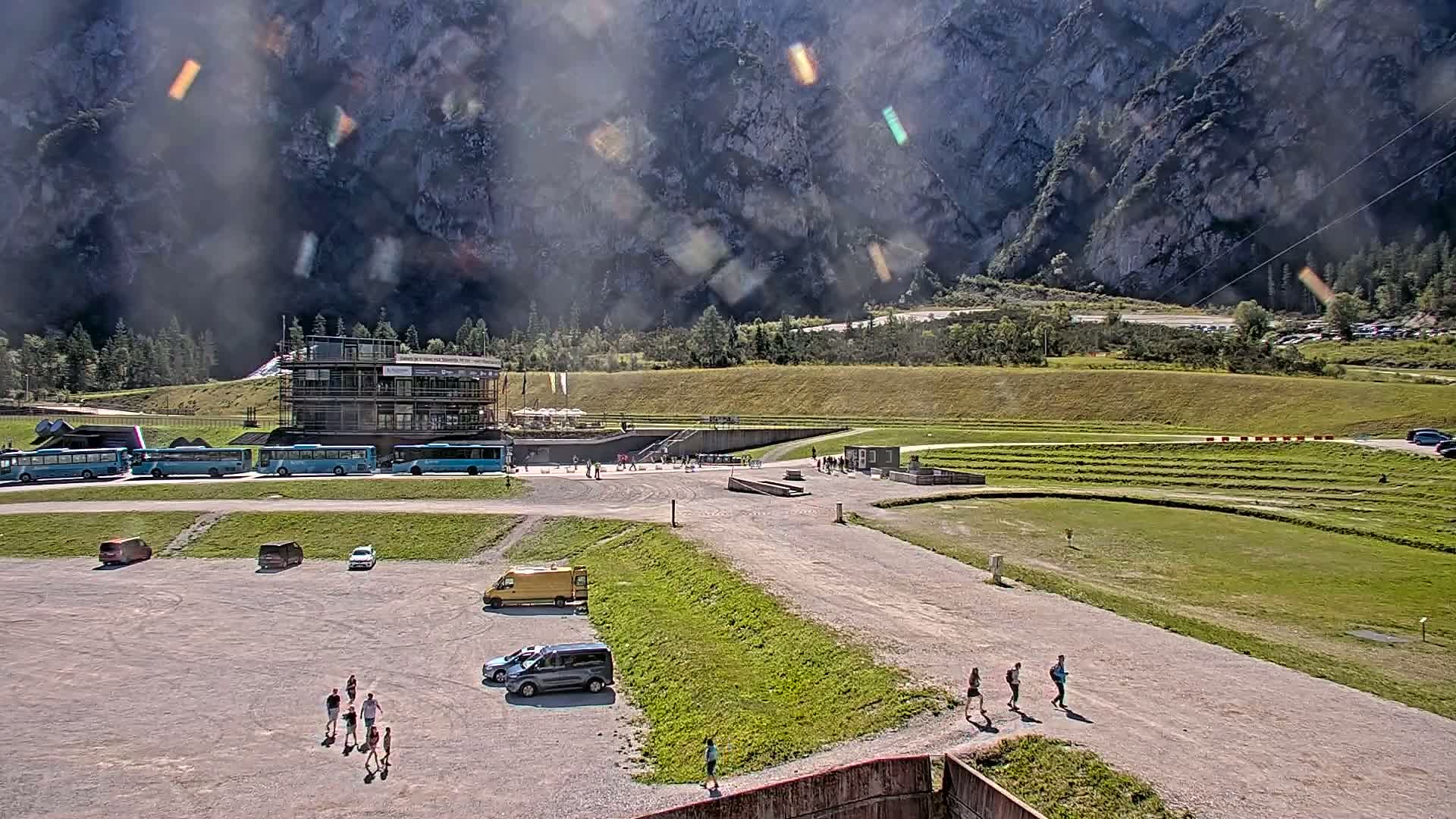 A parking lot with several vehicles and people walking, near a building and grassy area, with a large rocky mountain in the background under hazy, sunny conditions.