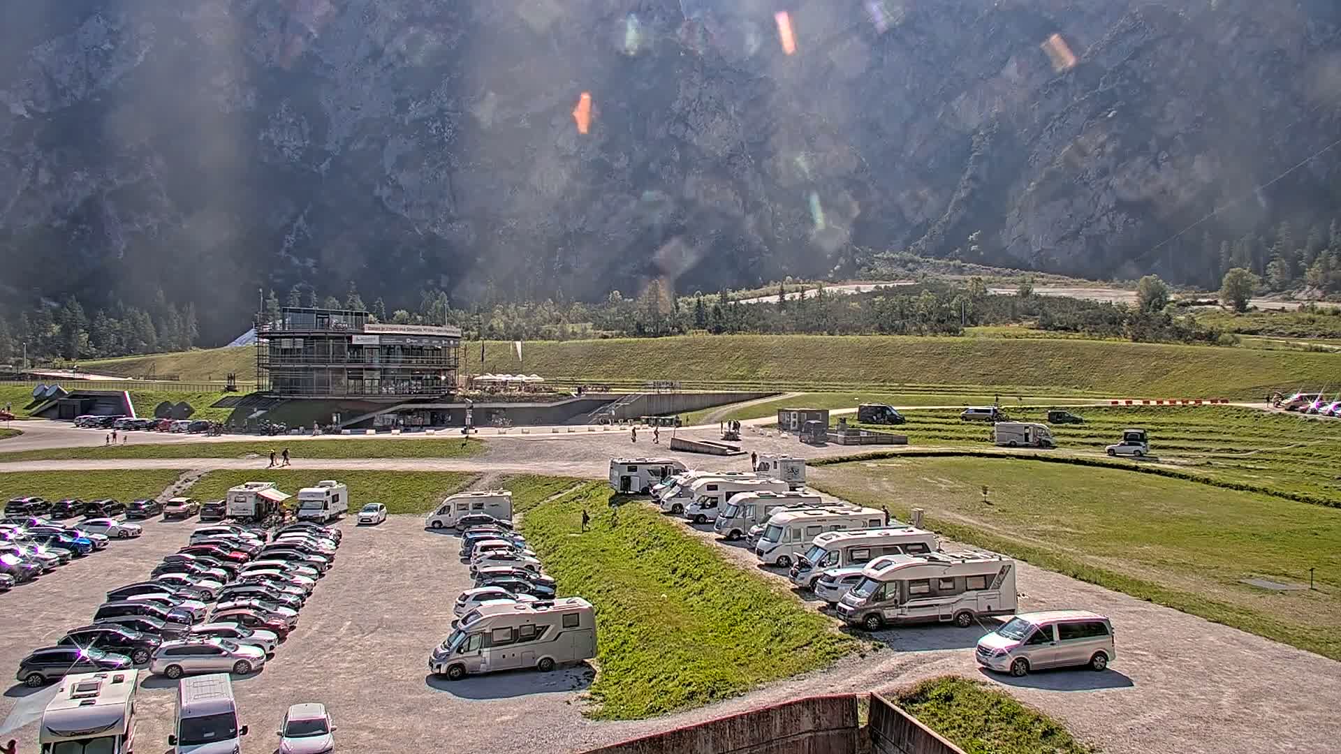 A large parking lot filled with cars and RVs is situated below a multi-story building, all set against a backdrop of mountains under a sunny sky.