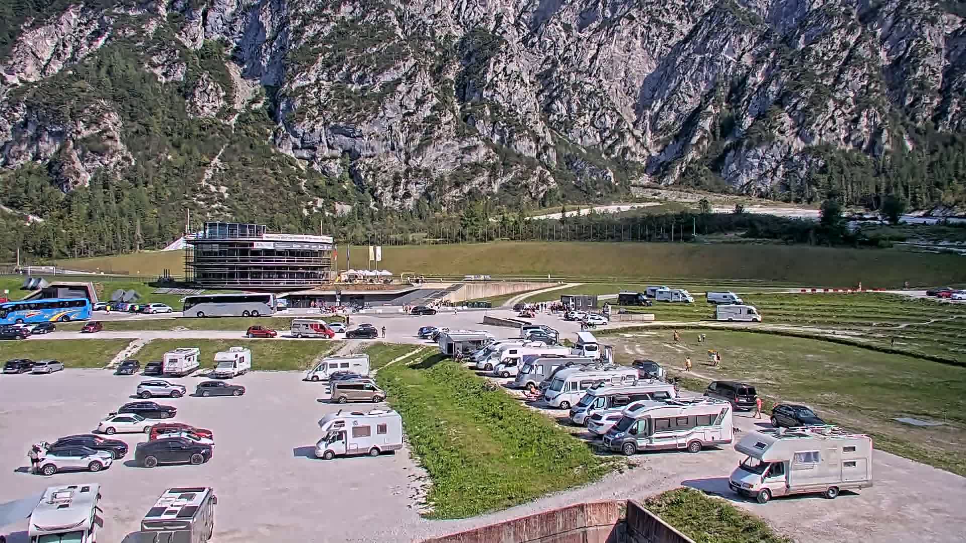 A large parking lot filled with cars and RVs is situated below a multi-story building, all set against a backdrop of mountains under a sunny sky.
