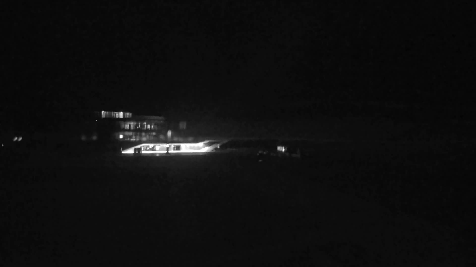 A very dark and blurry grayscale image taken at night reveals a distant building with glowing windows and a bright horizontal light source at its base.