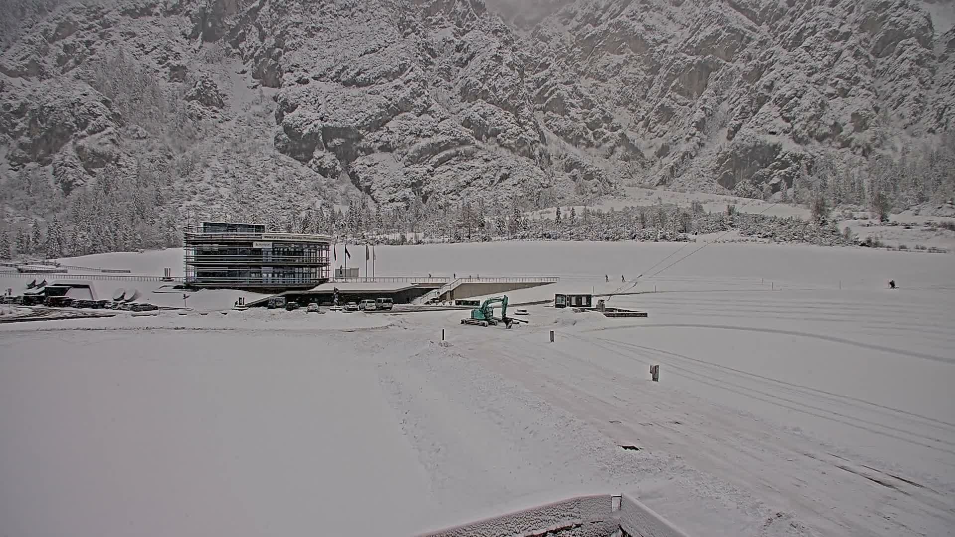 On an overcast winter day, a modern multi-story building, vehicles, and an excavator stand on a vast snow-covered plain or frozen lake, framed by majestic snow-dusted mountains.