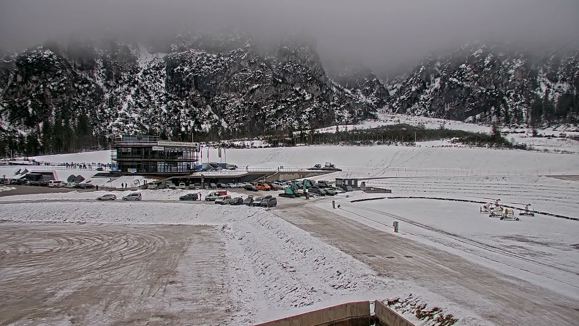 A snowy winter scene reveals a modern multi-story building surrounded by parked cars and an excavator, two small helicopters on a snow-dusted tarmac, and what appears to be a bobsled track, all set against a backdrop of snow-covered mountains partially obscured by thick, gray overcast clouds.