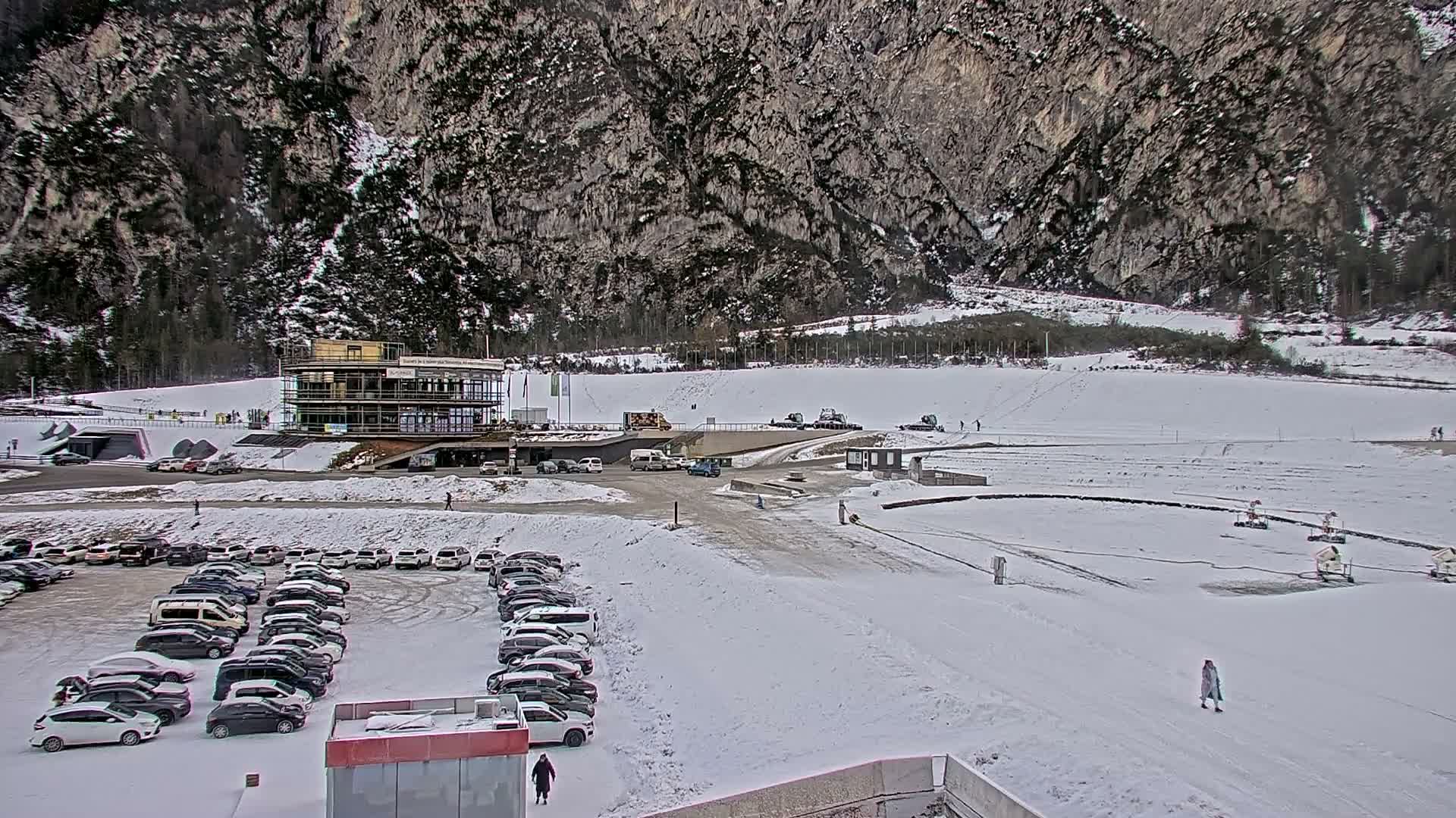 A snowy winter scene reveals a modern multi-story building surrounded by parked cars and an excavator, two small helicopters on a snow-dusted tarmac, and what appears to be a bobsled track, all set against a backdrop of snow-covered mountains partially obscured by thick, gray overcast clouds.