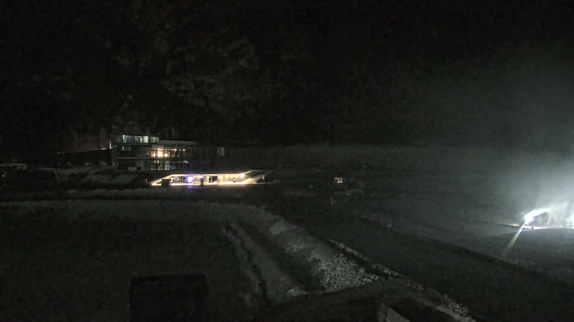A snowy winter scene reveals a modern multi-story building surrounded by parked cars and an excavator, two small helicopters on a snow-dusted tarmac, and what appears to be a bobsled track, all set against a backdrop of snow-covered mountains partially obscured by thick, gray overcast clouds.